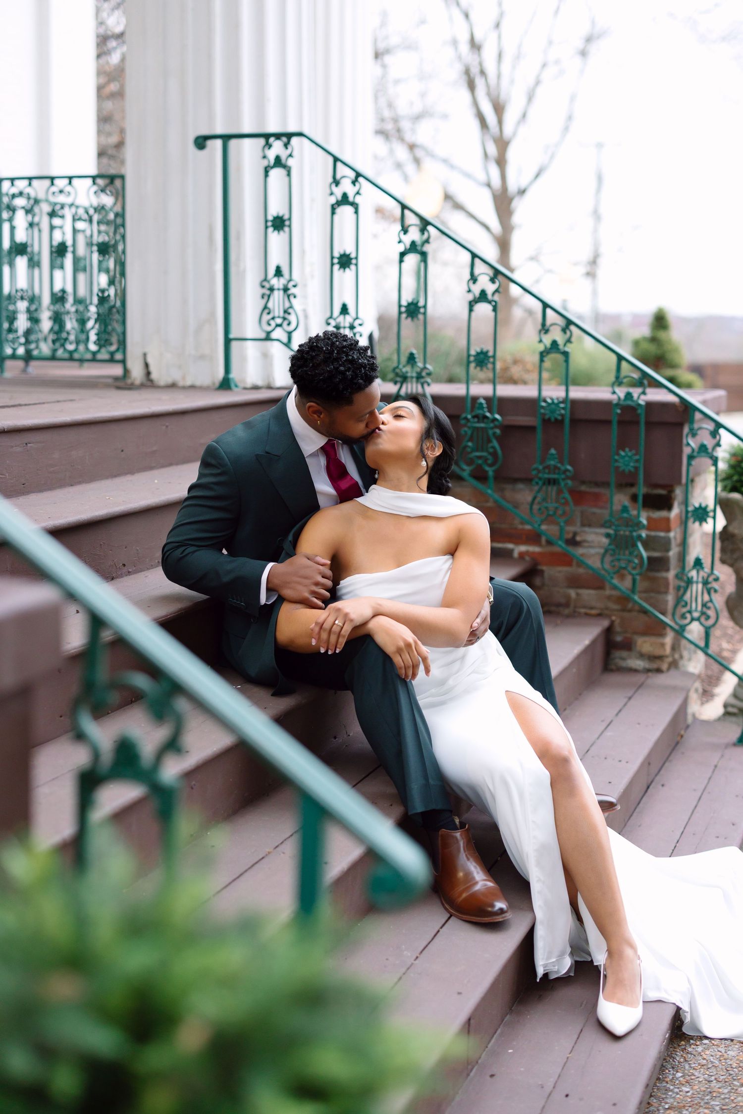 Bride and groom share a tender kiss on decorated steps, she wears a white halter gown, he wears a dark green suit.