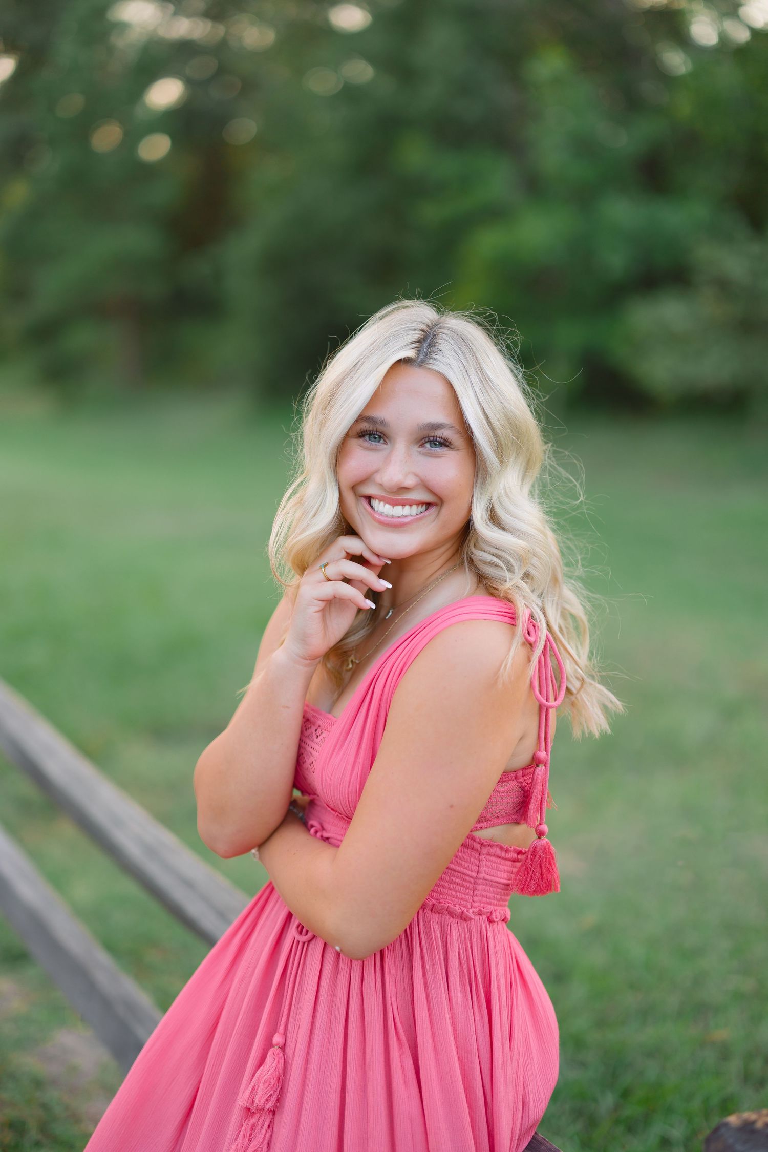 A person in a coral pink summer dress poses playfully in a grassy outdoor setting.