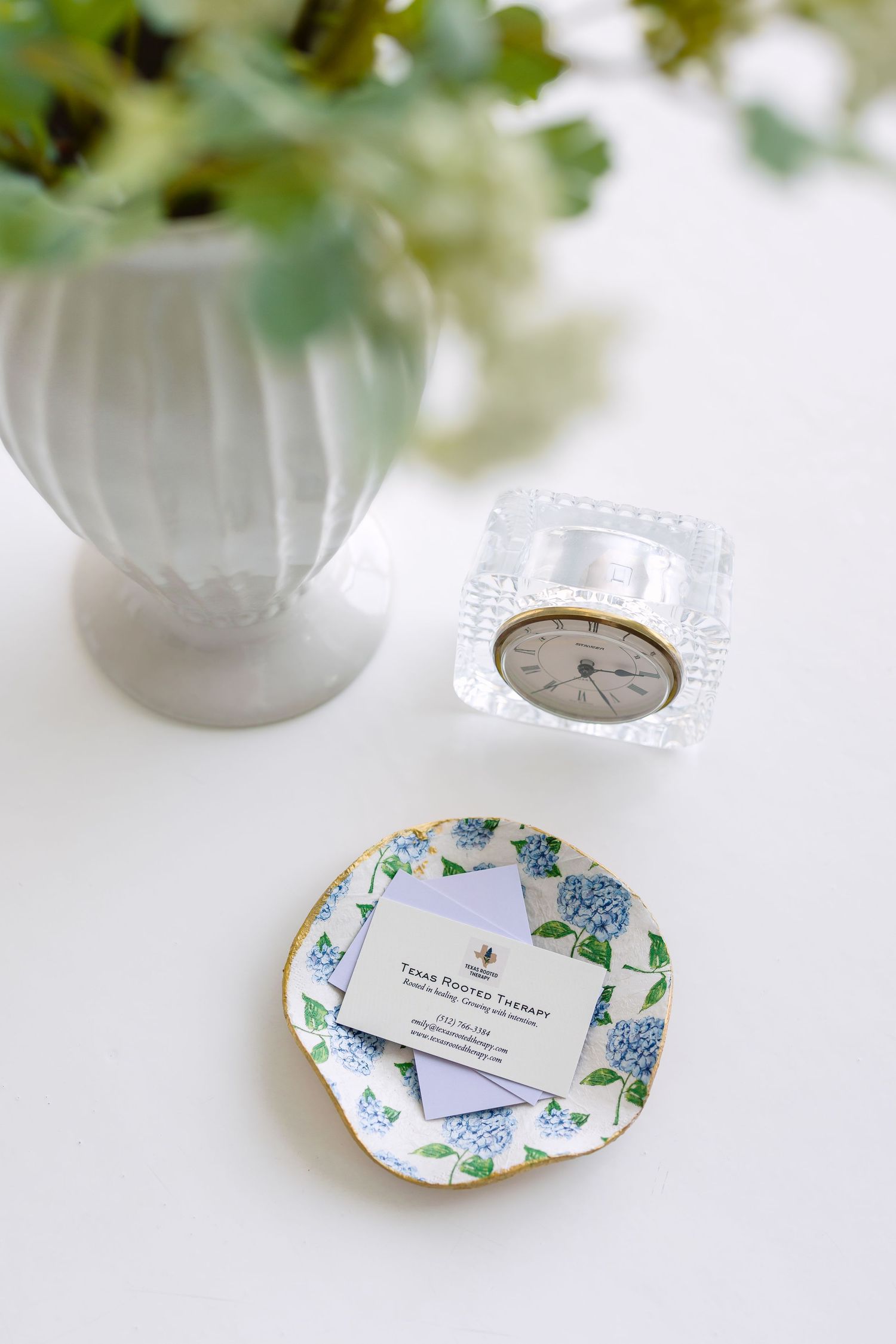 Elegant desk vignette with floral business cards on a decorative plate, crystal clock, and white vase with greenery.