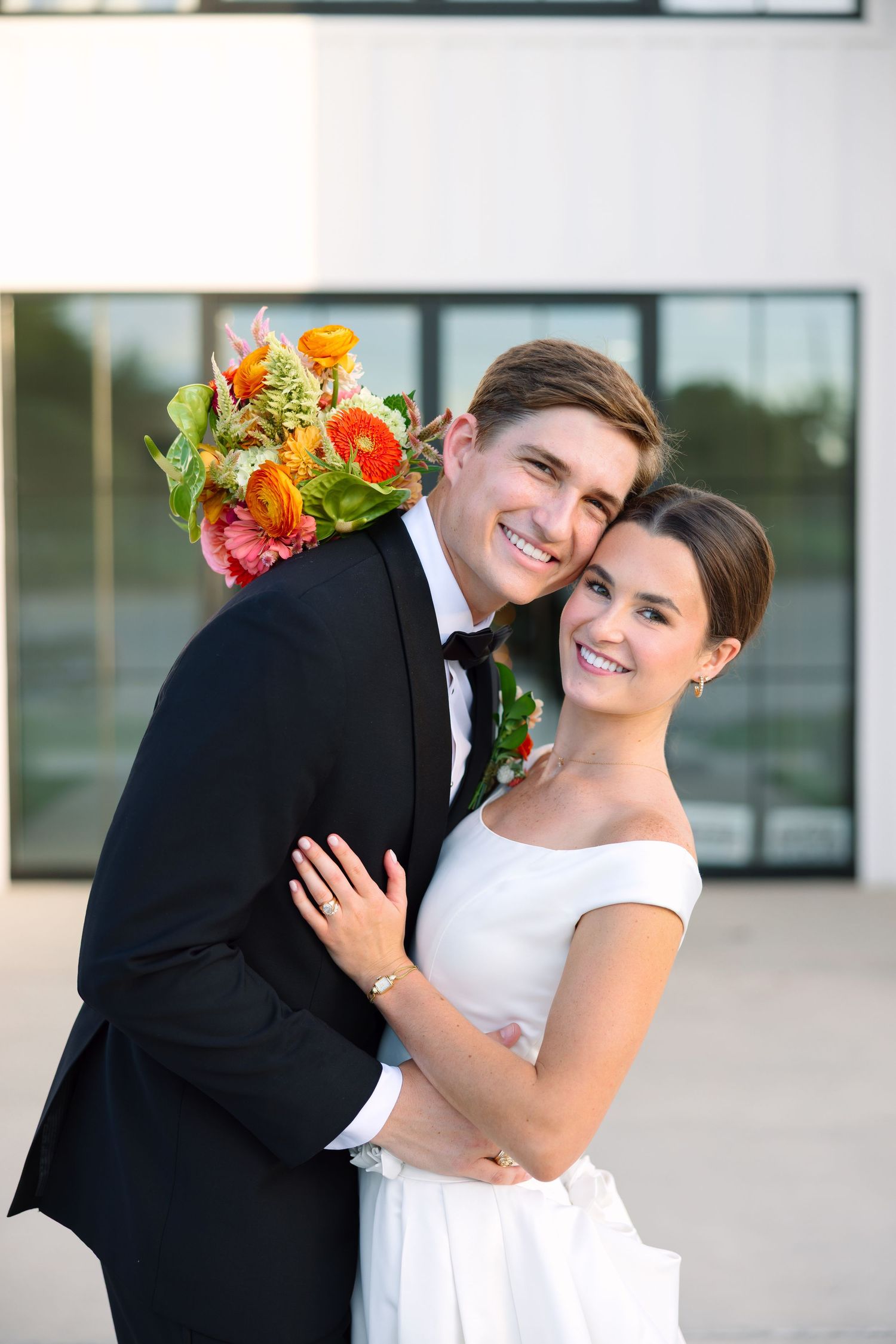 Smiling bride and groom embracing outdoors, groom in black tuxedo holding colorful bouquet, bride in white off-shoulder gown.