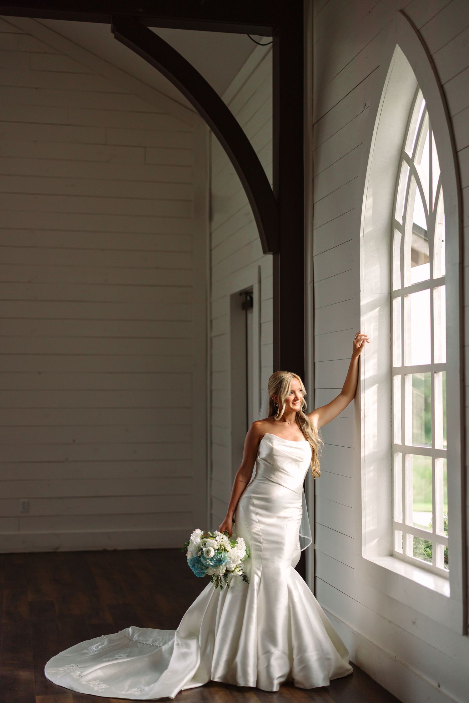 Bride in flowing white gown poses gracefully near arched church windows.