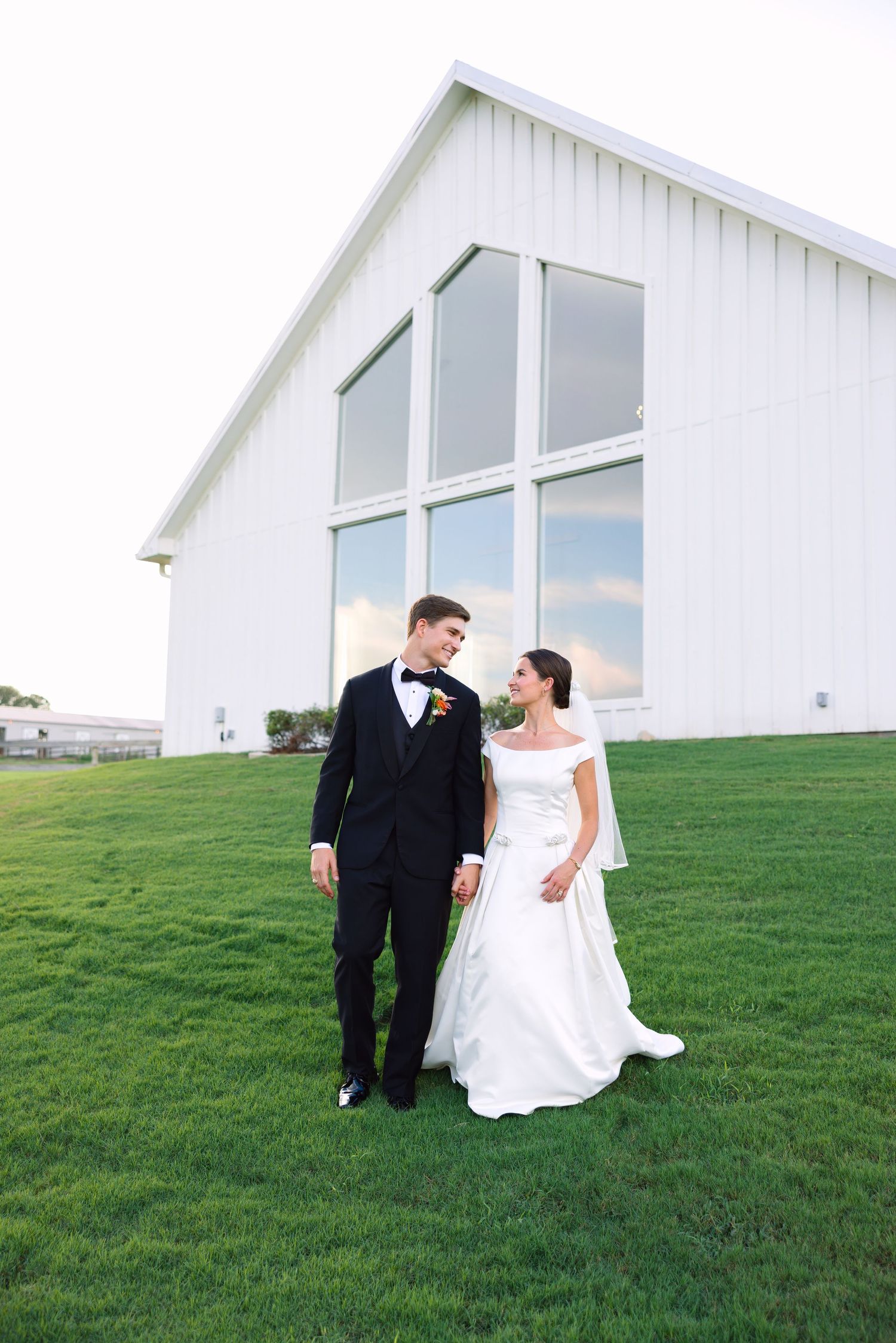 Bride and groom walking hand in hand on green lawn in front of white barn wedding venue.