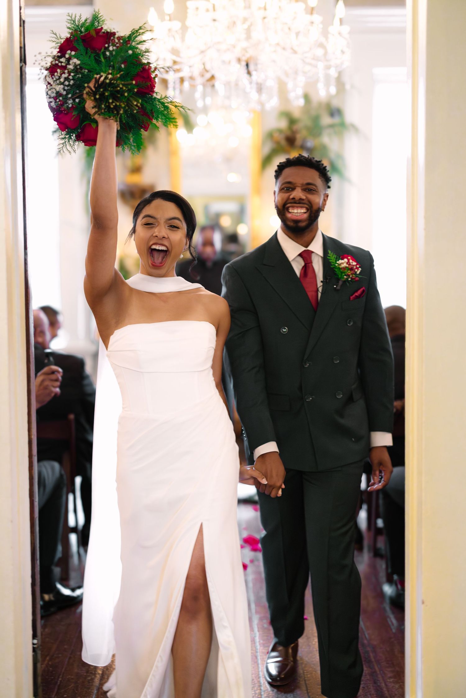 Joyful newlyweds enter reception hall as bride raises red bouquet and groom smiles in dark green suit.