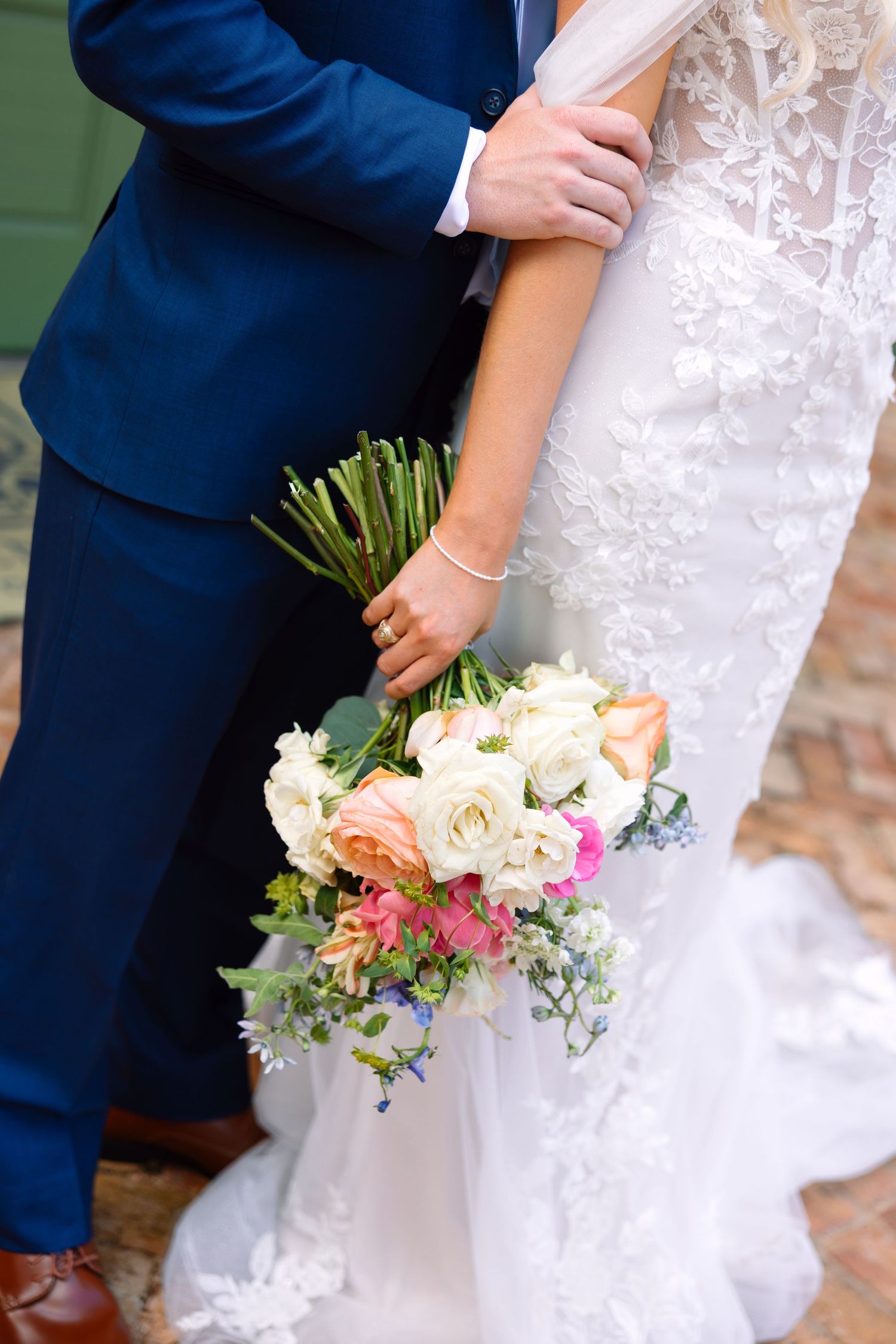 Close-up of bride in lace gown holding colorful wedding bouquet with groom in navy suit.