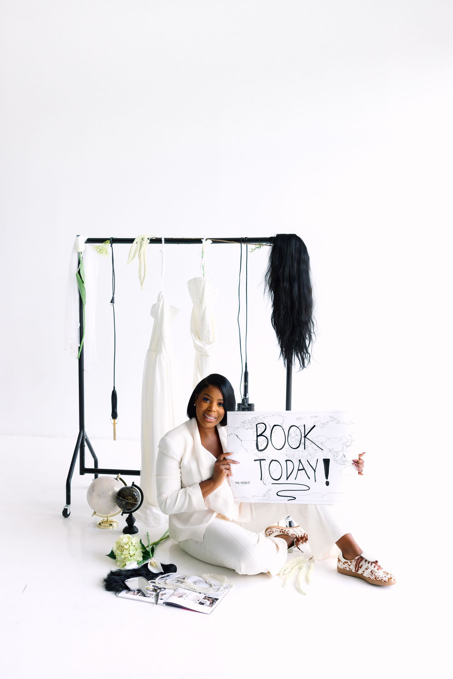 A smiling woman in white sits on the floor holding a Book Today sign near a clothing rack with accessories.