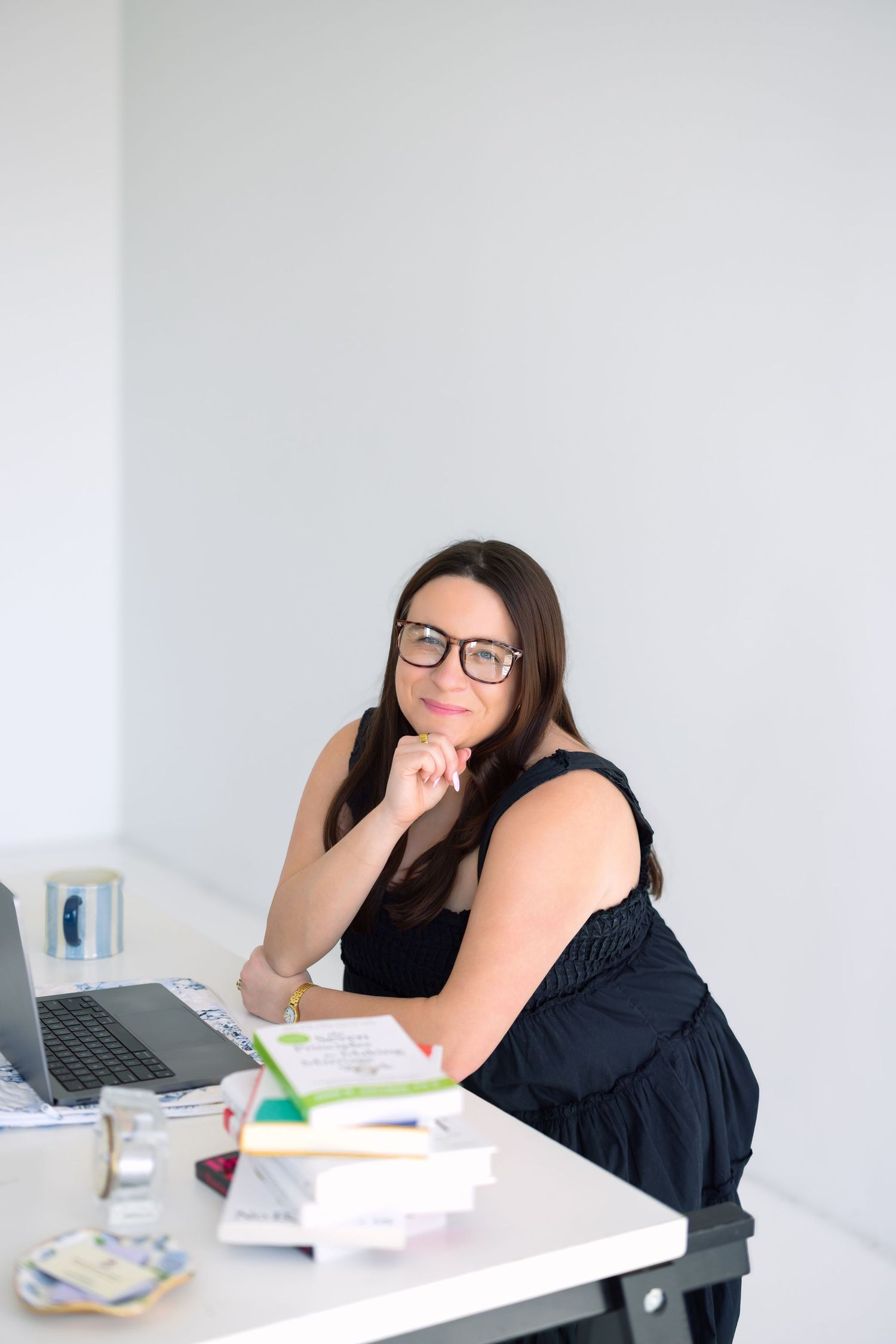 Woman in glasses sitting at desk with laptop and books, resting chin on hand in bright office.