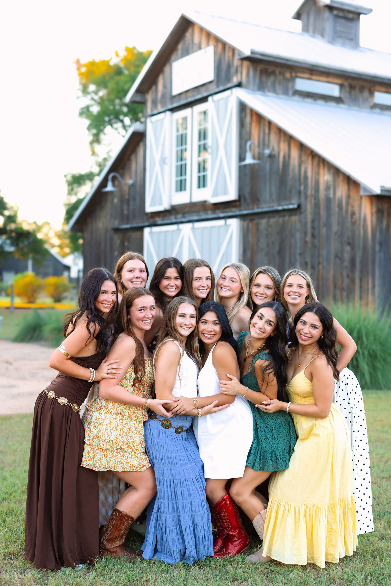 Friends in colorful dresses embrace in a group hug outside a weathered barn.