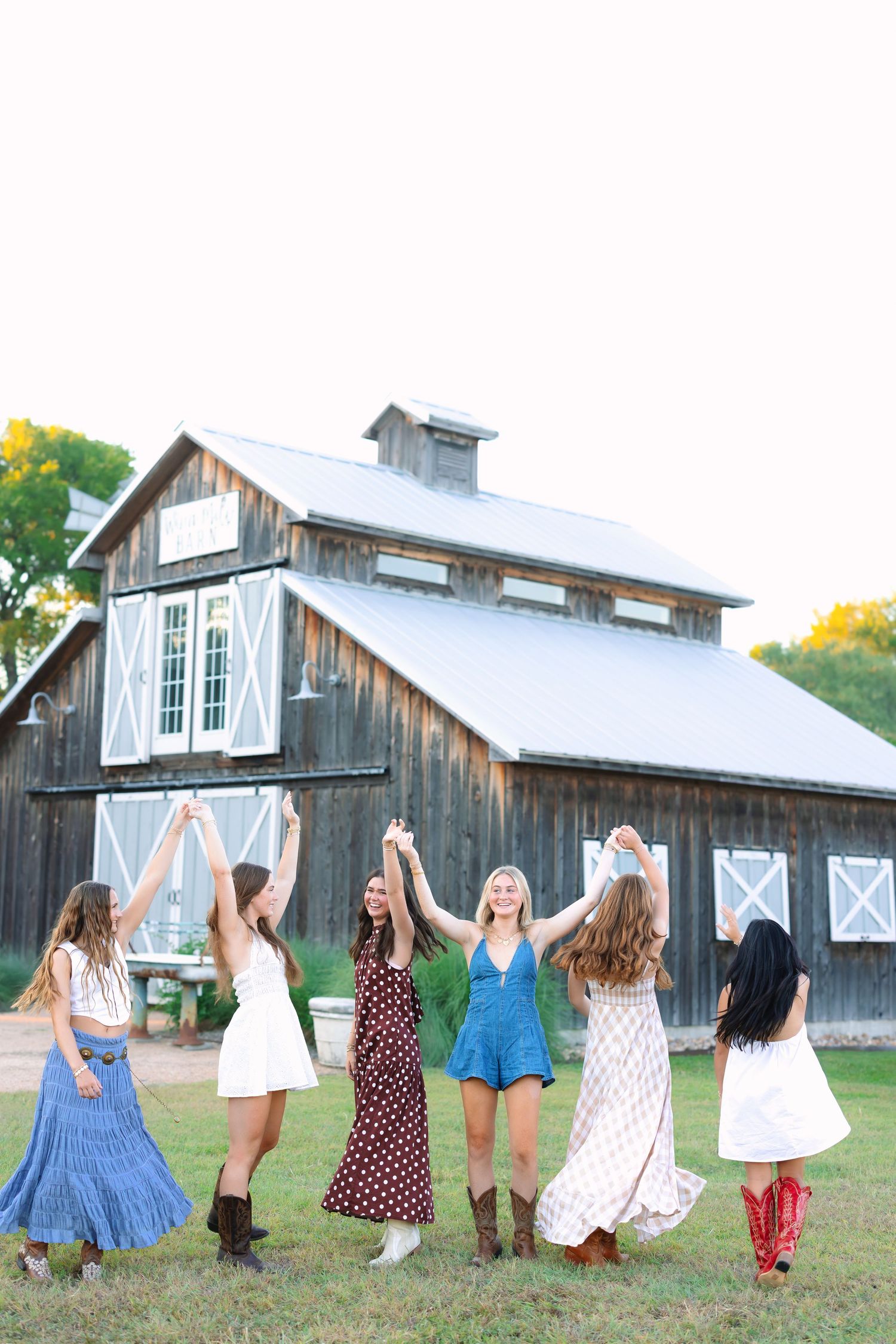 Friends in summer dresses and boots jumping joyfully in front of a rustic barn.