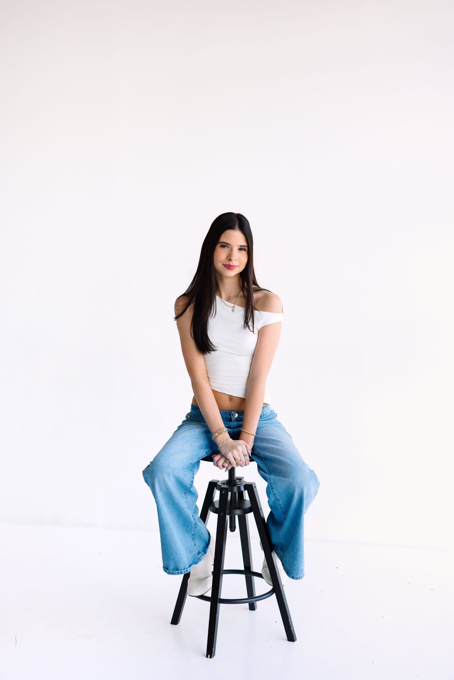 Casual studio portrait sitting on black stool wearing white top and light blue jeans.
