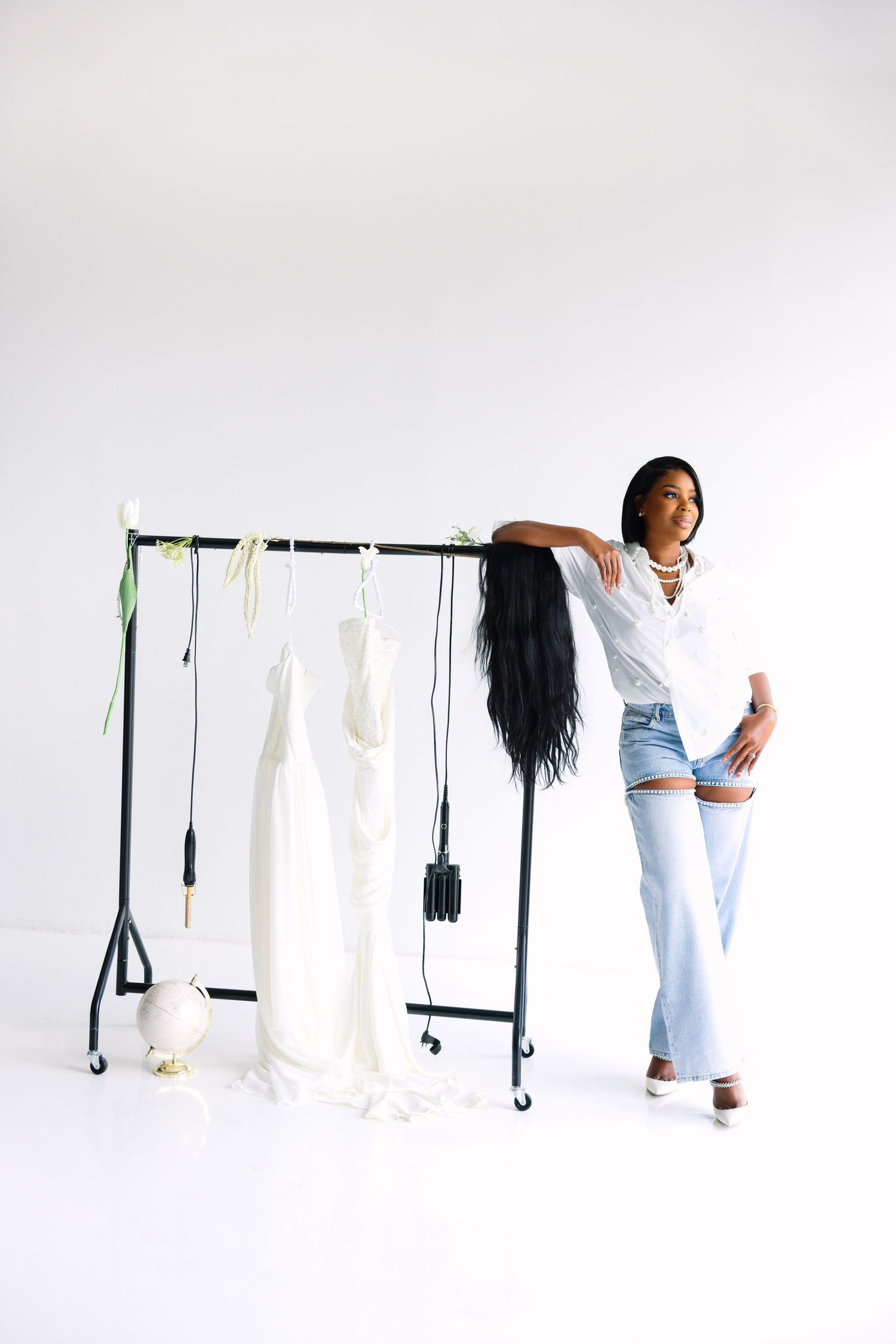 Stylish woman in white shirt and jeans leans against black clothing rack with white dresses in studio.