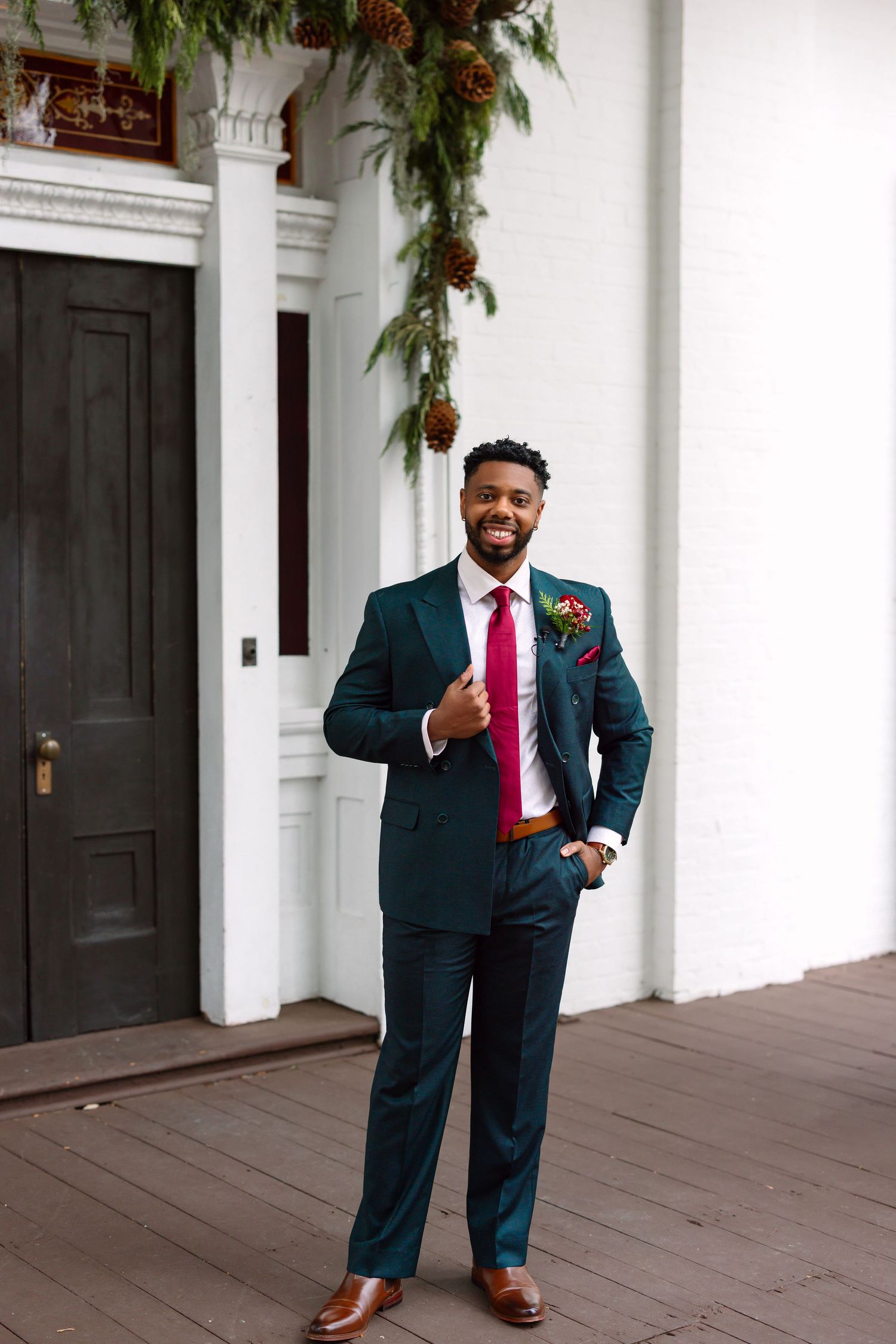 Smiling groom in teal suit, red tie, boutonniere, brown shoes, standing by white building with pine cone garland.
