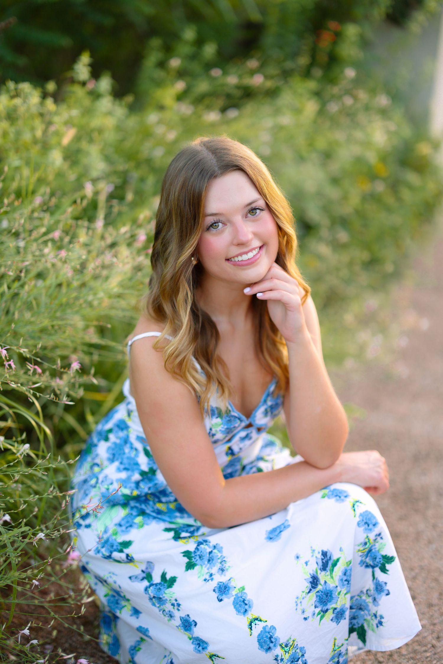 Young woman in white floral blue dress sitting outdoors among green plants, smiling warmly with hand near chin.