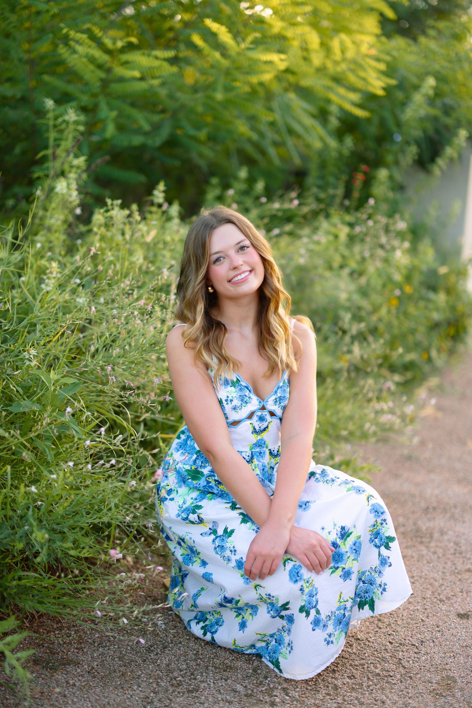 Young woman in white floral dress sitting outdoors among green plants, smiling warmly at camera.