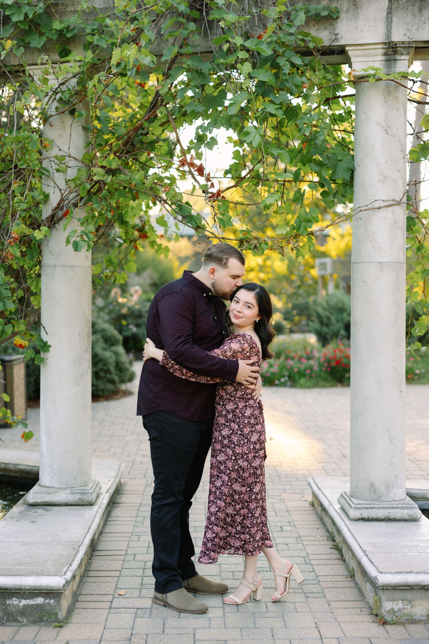 Couple embracing and kissing under a vine-covered pergola with white columns in a romantic garden.