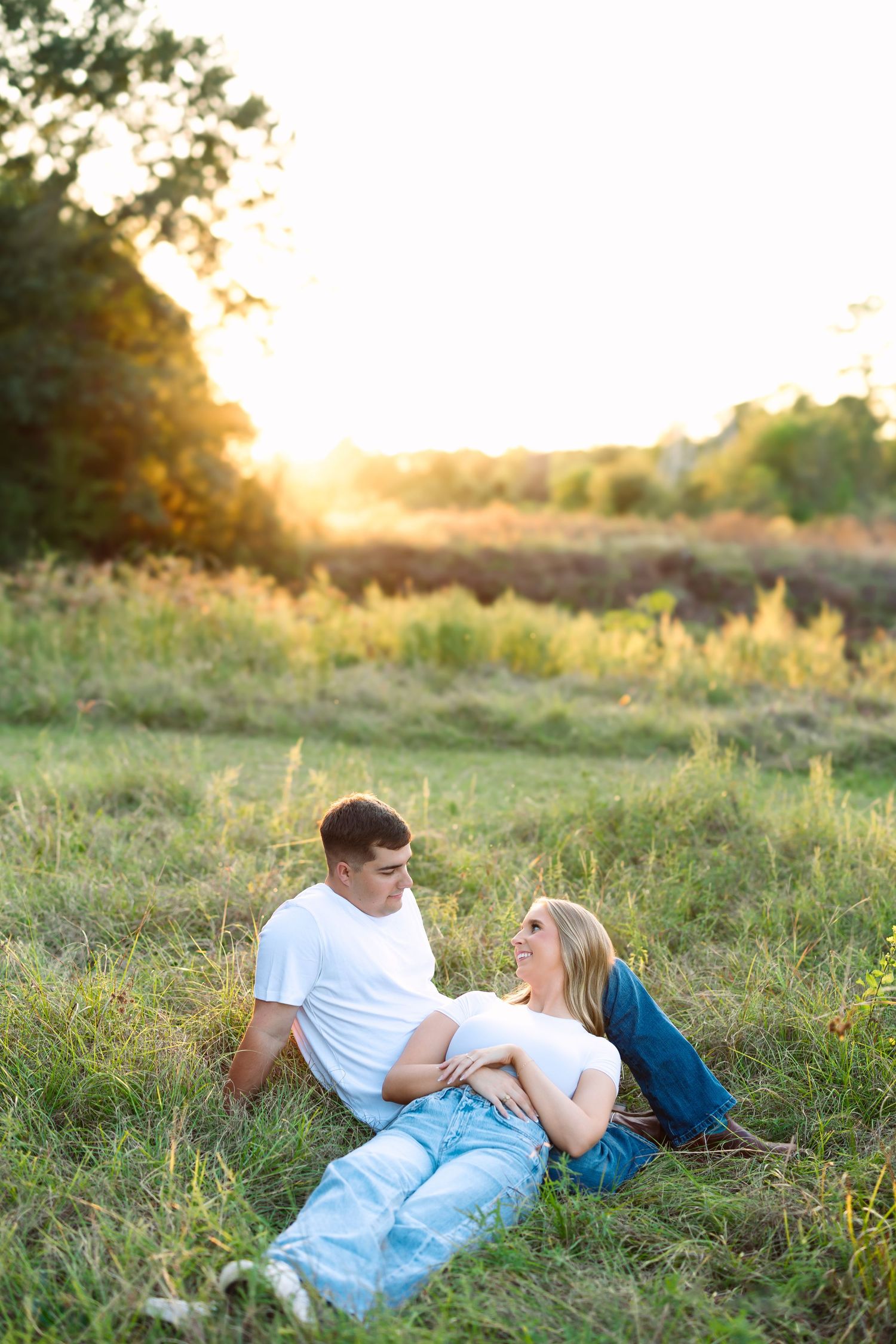 A couple relaxes in a sunlit grassy field at golden hour, the woman lying in the man's lap as they gaze at each other.