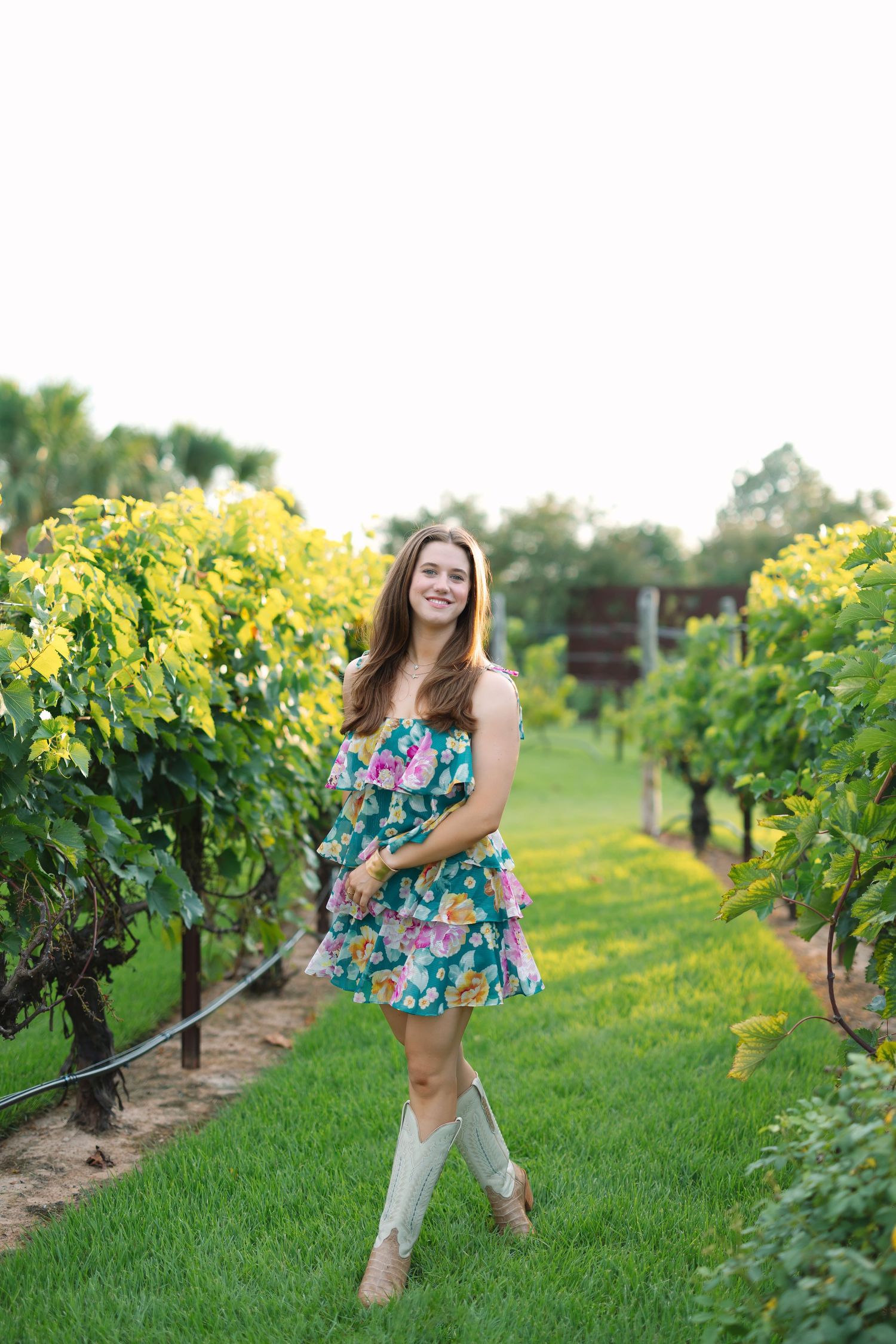 Woman in floral dress poses playfully in vineyard during sunset.