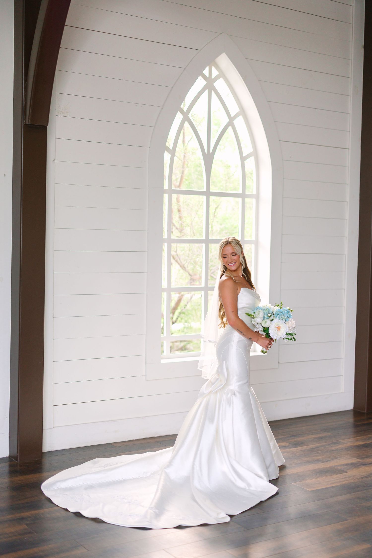 A bride in a strapless gown stands in an elegant white chapel with gothic windows.