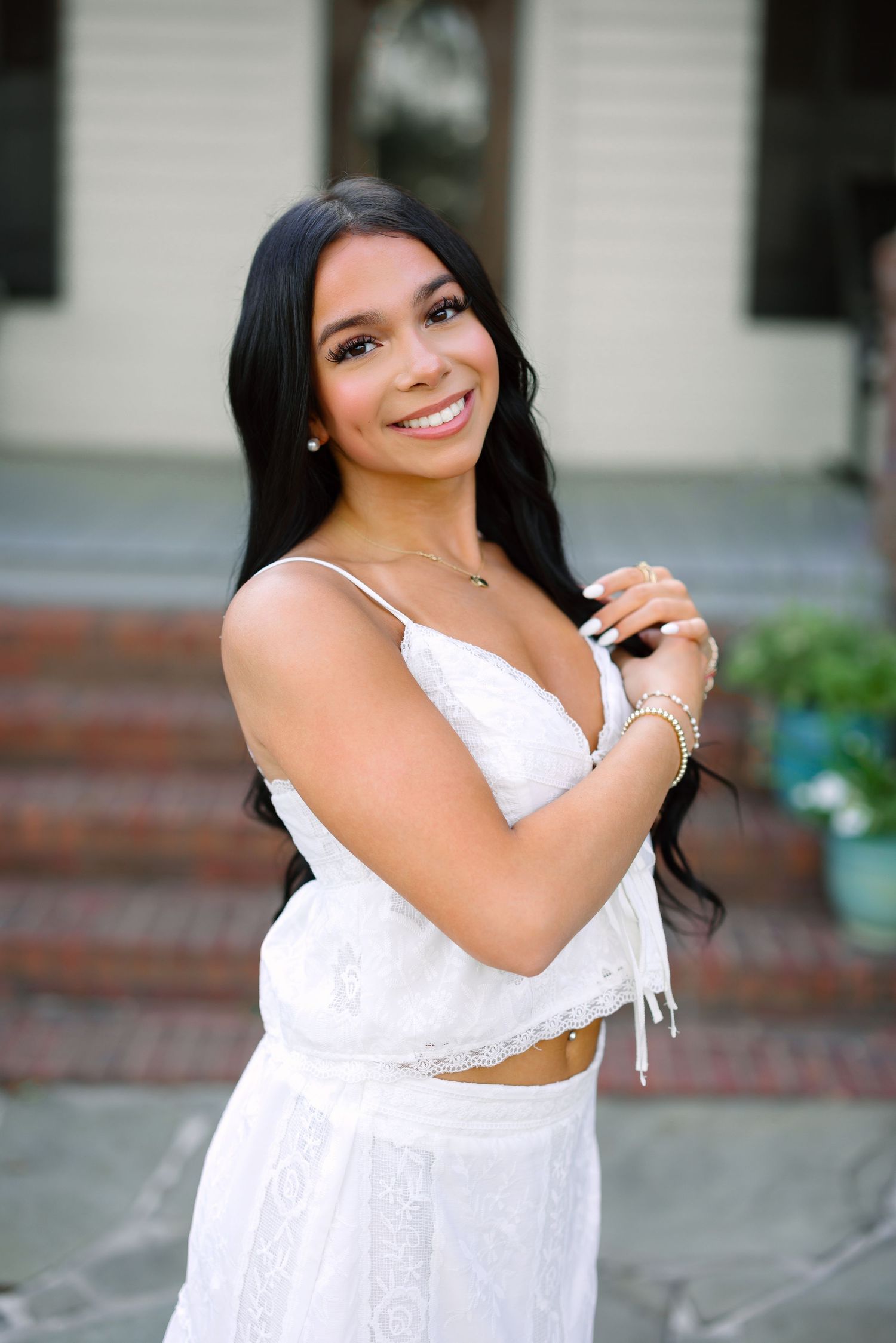 Young woman in white lace dress smiling outdoors, long dark hair, gold jewelry, brick steps background.