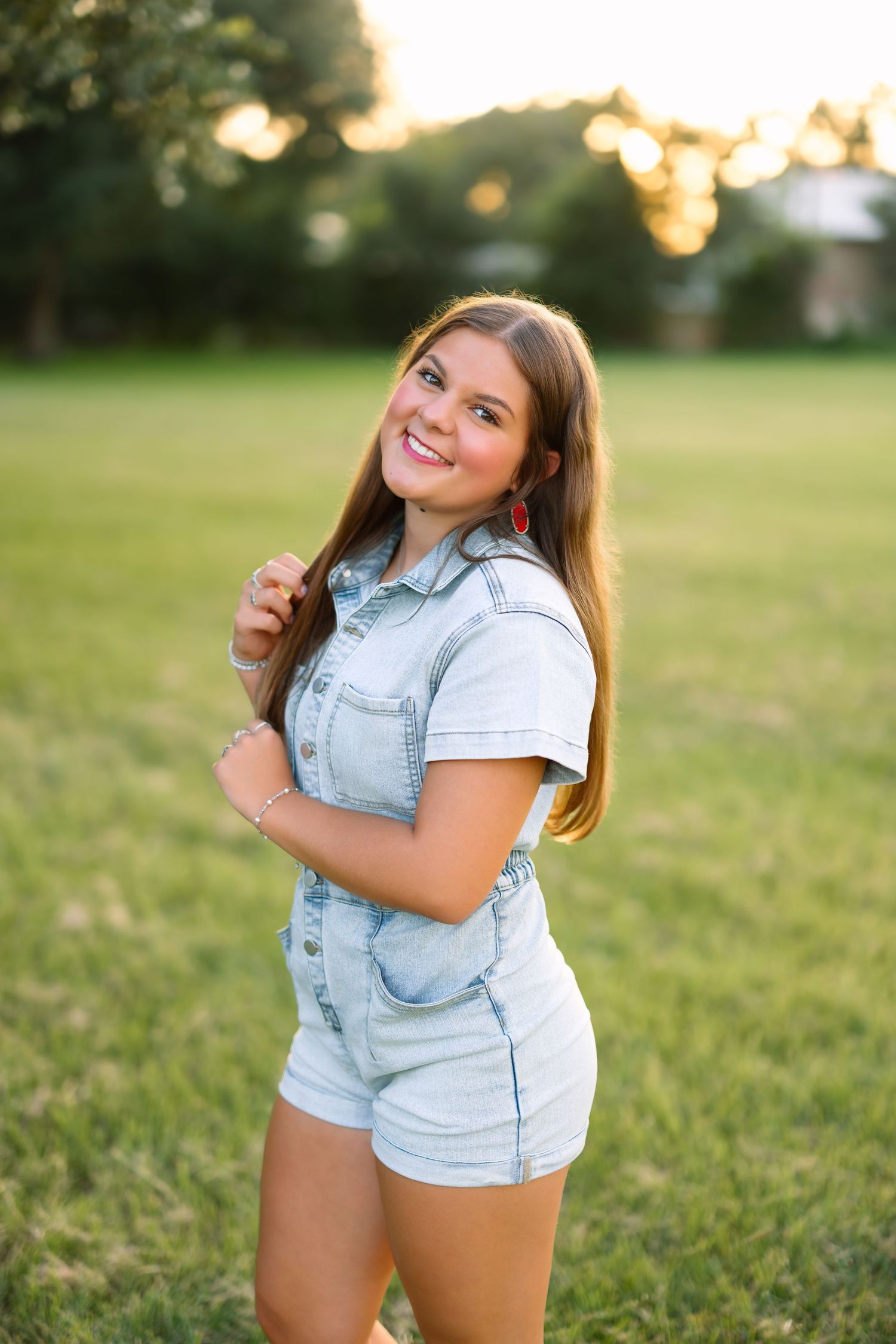 Smiling teenage girl in denim romper poses outdoors on green grass at golden hour sunset.