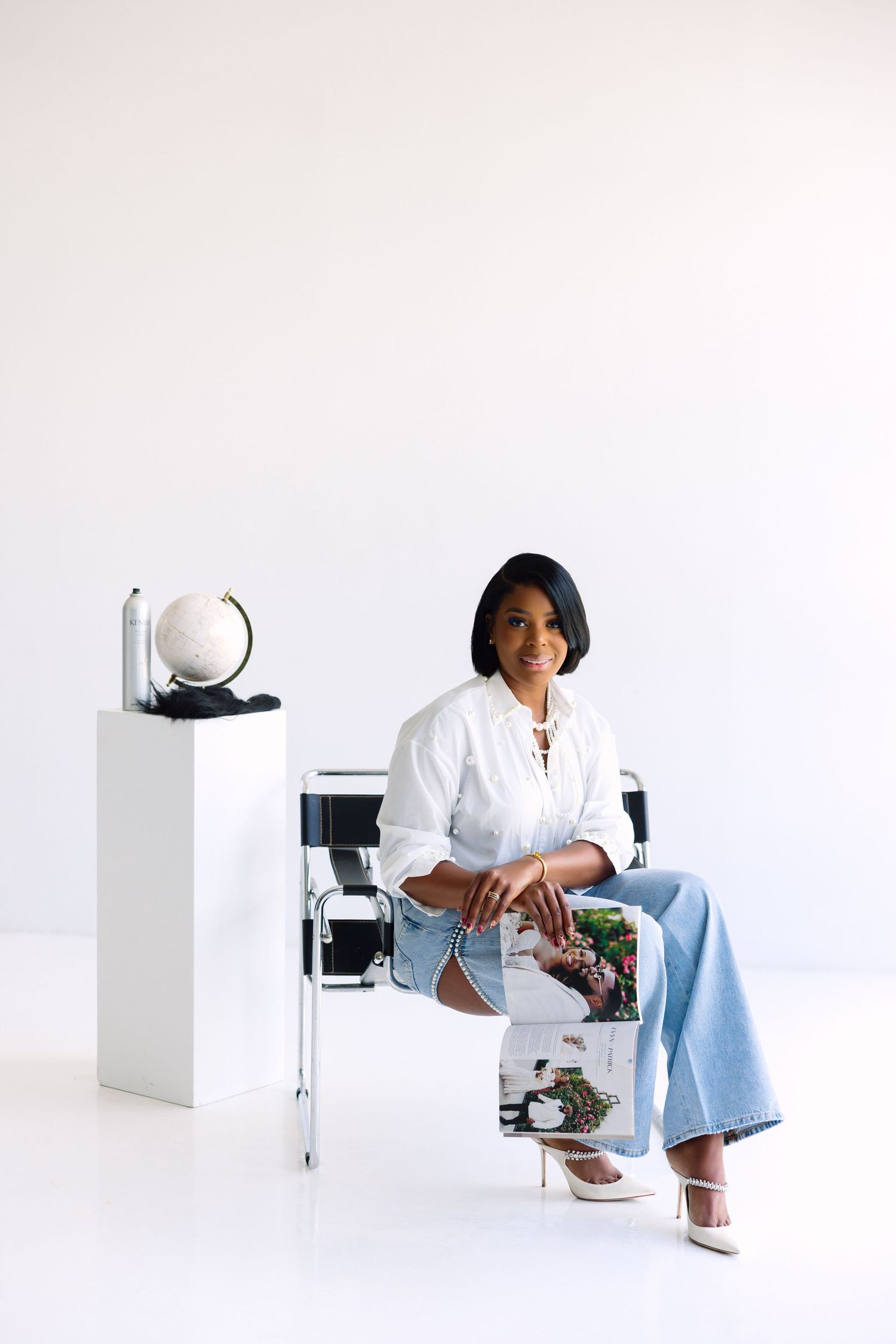 A confident Black woman sits in a modern chair wearing a white blouse and jeans, holding a magazine beside a globe display.