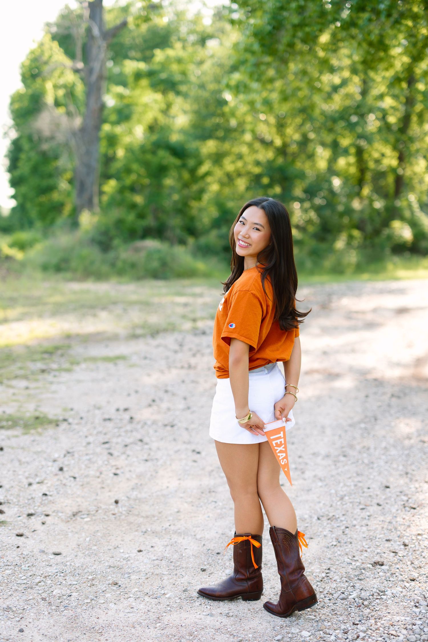 Young woman in orange top, white skirt, and cowboy boots smiles on a gravel path surrounded by green trees.