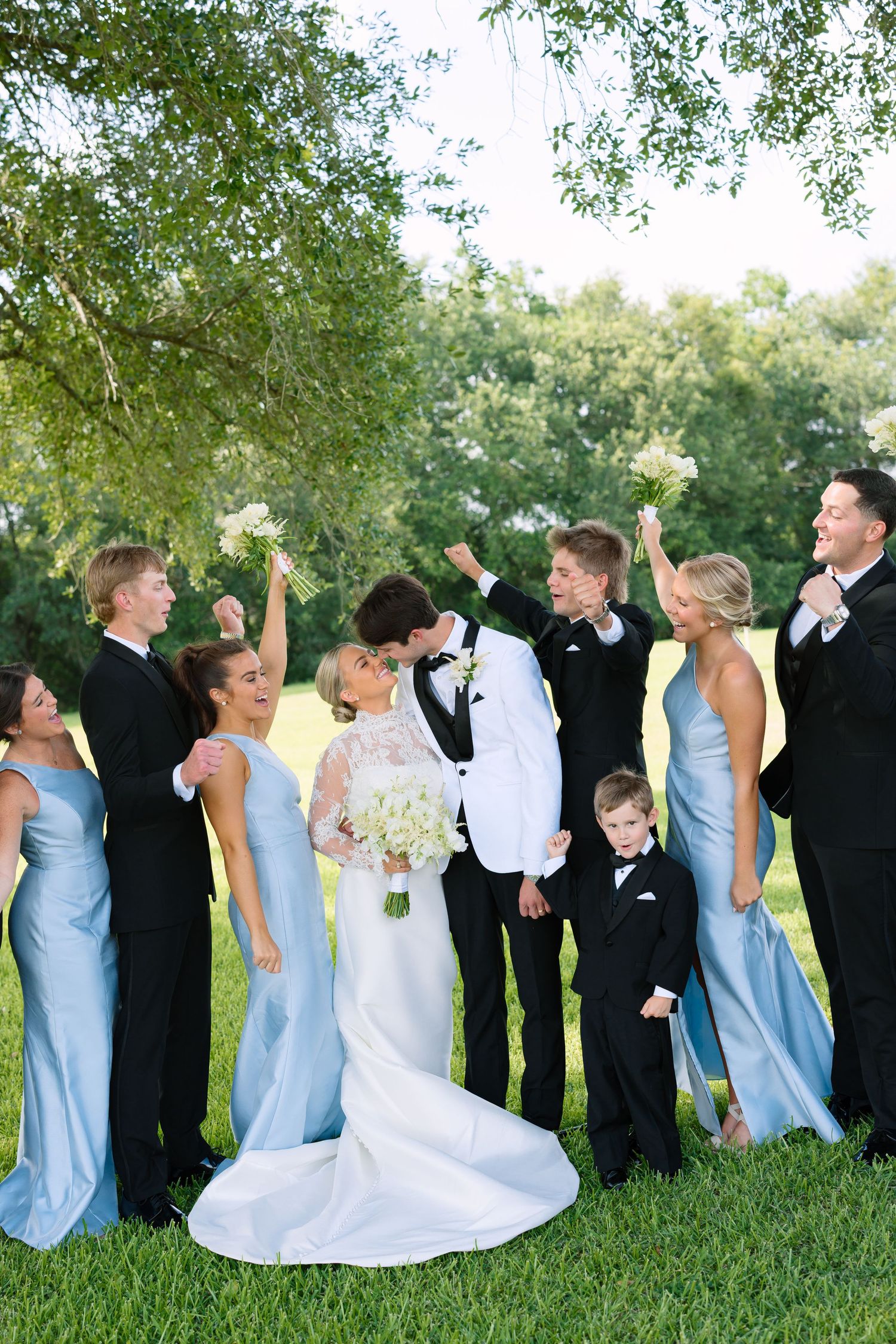 Wedding party in black tuxedos and light blue gowns cheering as bride and groom share a kiss outdoors.