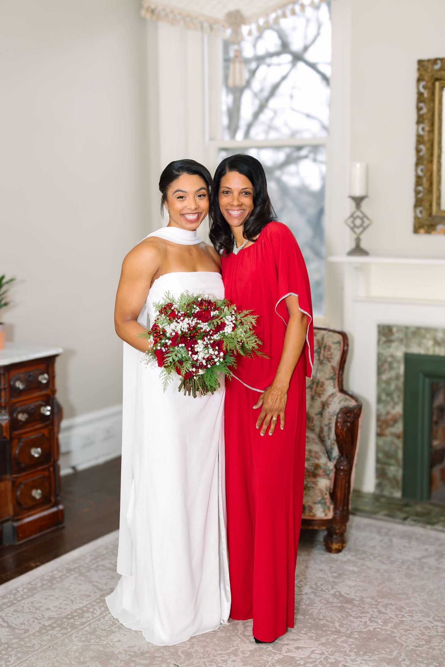 Bride in white gown holding red bouquet poses with woman in red dress indoors near fireplace.
