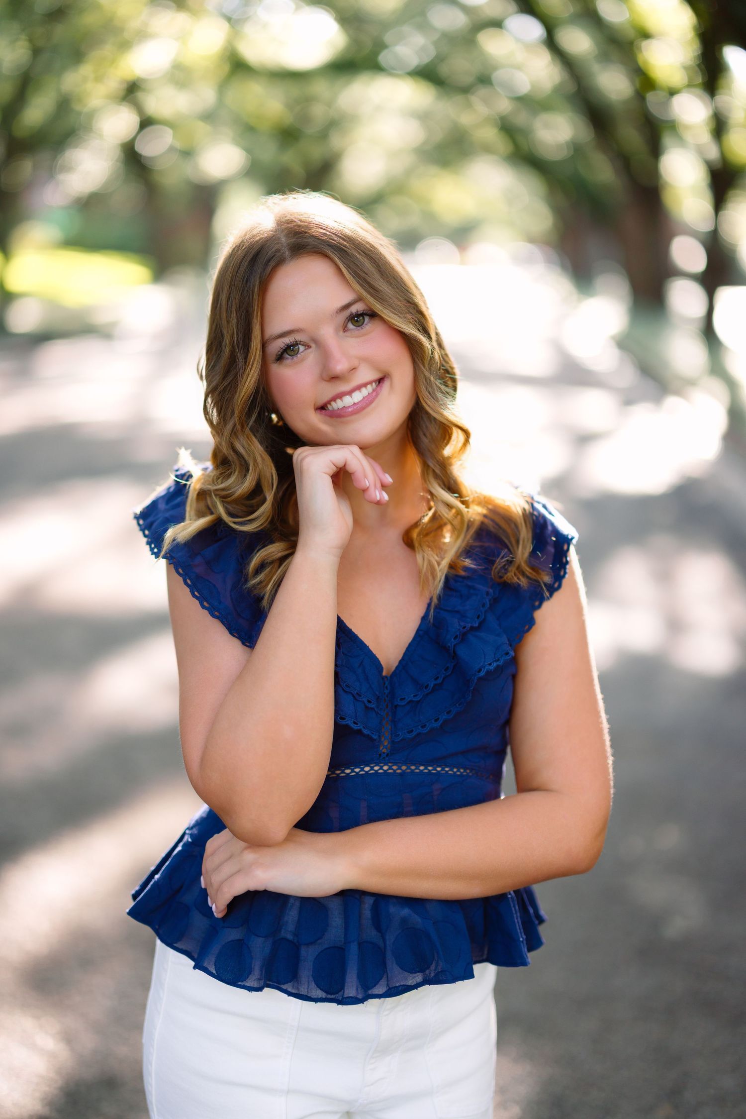 Young woman in blue ruffled top and white pants smiling outdoors in a tree-lined path.