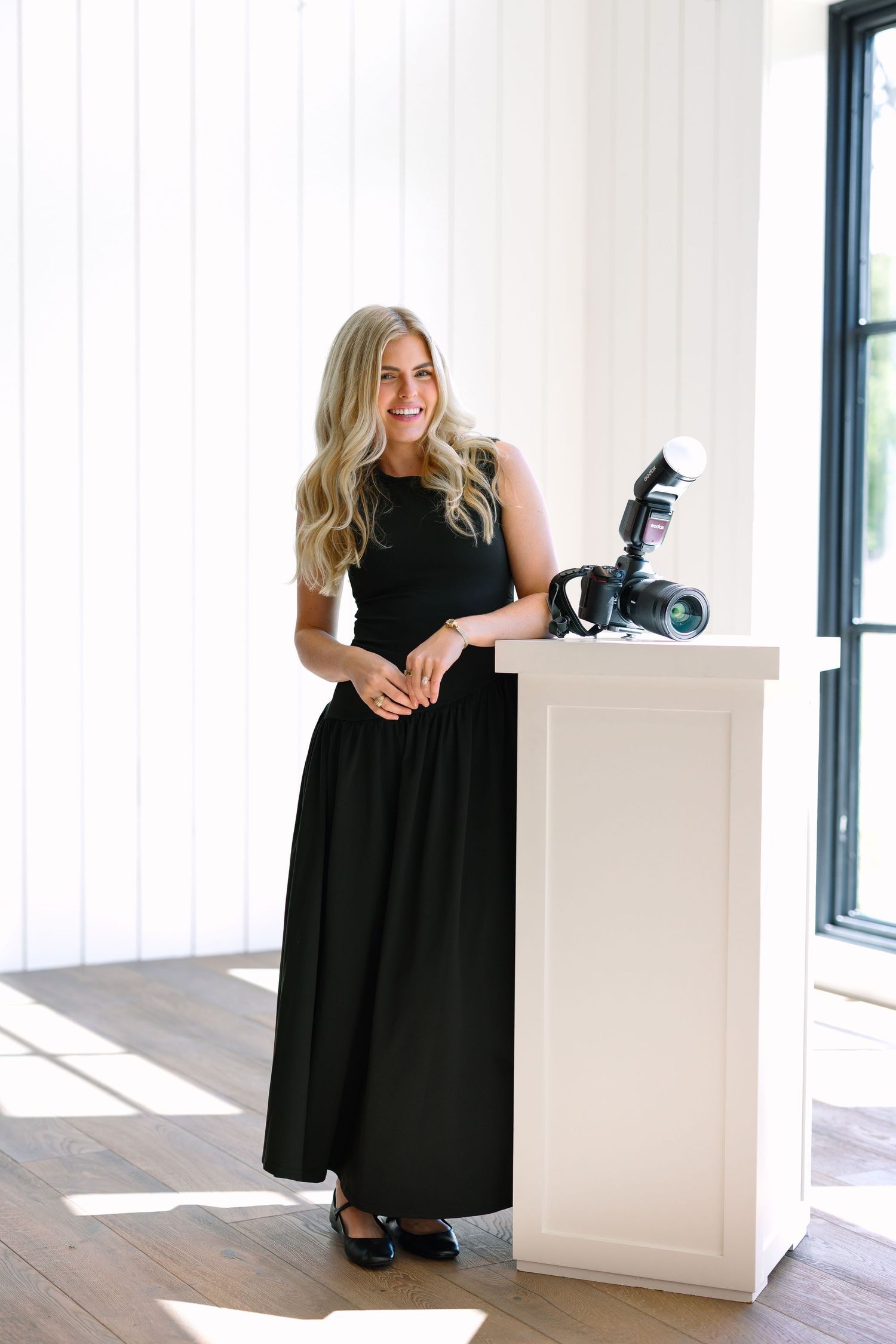 A smiling blonde woman in a black maxi dress stands beside a white pedestal with a camera in a bright studio.