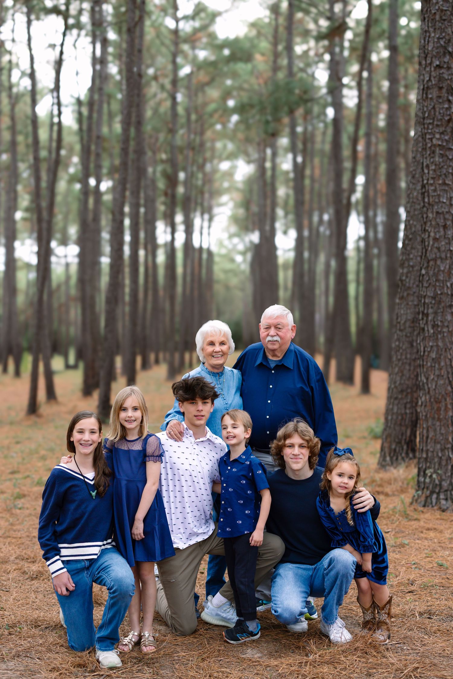 Multi-generational family portrait with grandparents and five grandchildren posing in a pine forest wearing blue outfits.