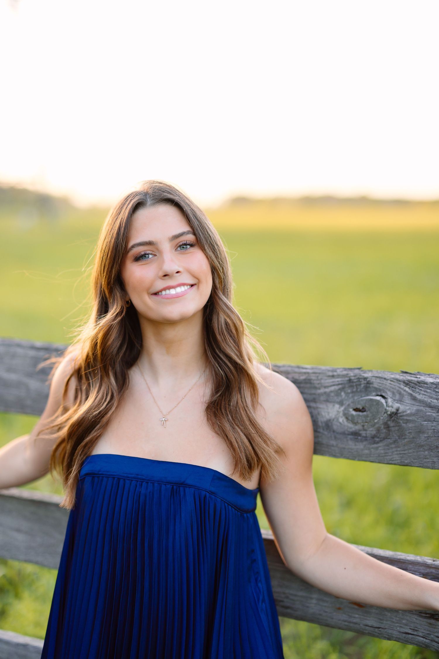 Senior portrait session featuring a royal blue off-shoulder dress against a rustic wooden fence and green field background.