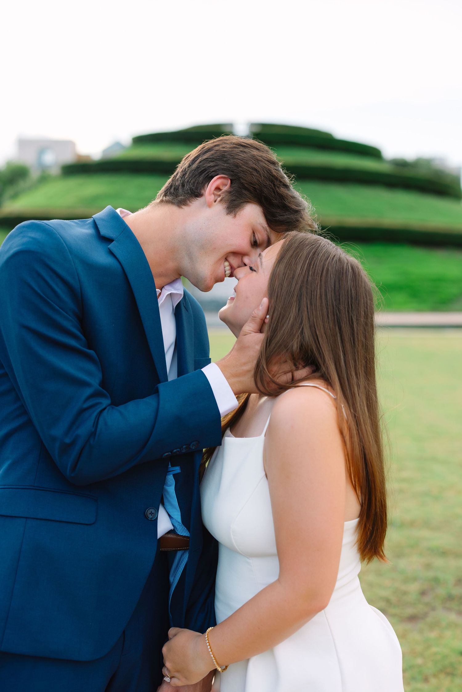 A tender moment between a couple in blue suit and white dress against a green hillside backdrop.