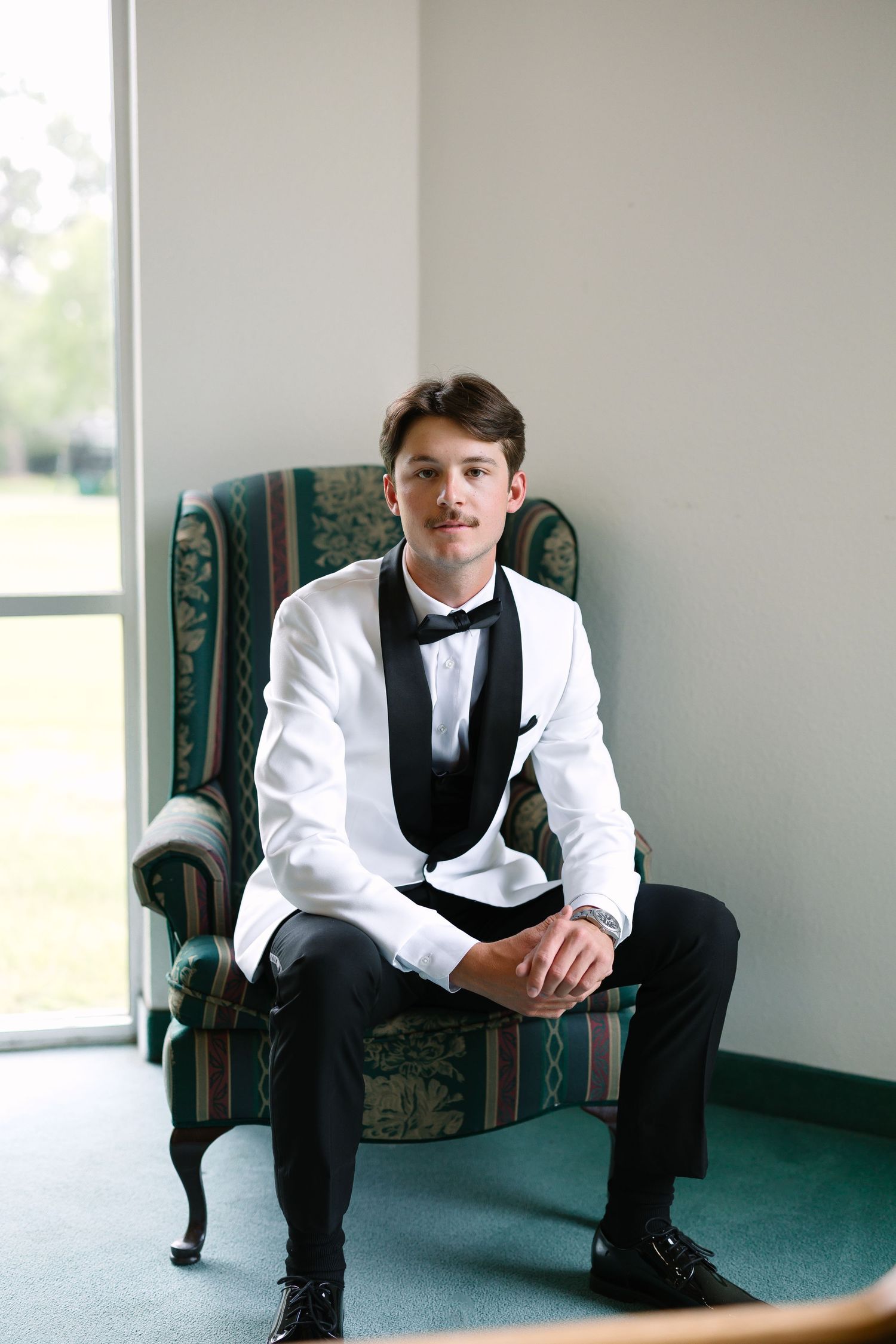 Groom in white tuxedo jacket with black lapels and bow tie seated in ornate green chair by window.