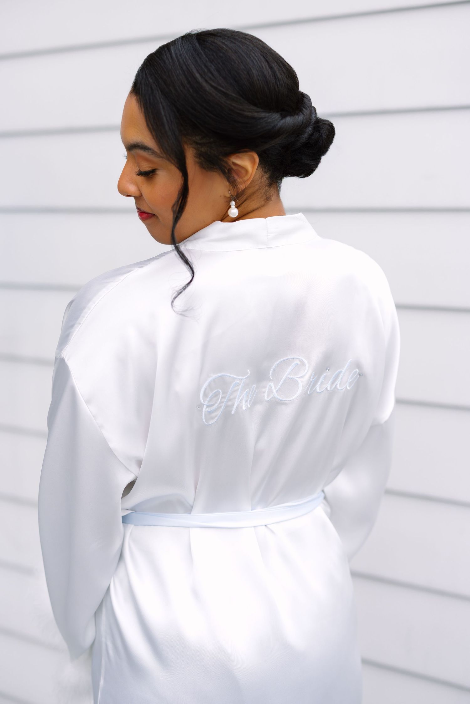 Bride wearing white satin robe with The Bride embroidery, showing elegant updo hairstyle and pearl earring.