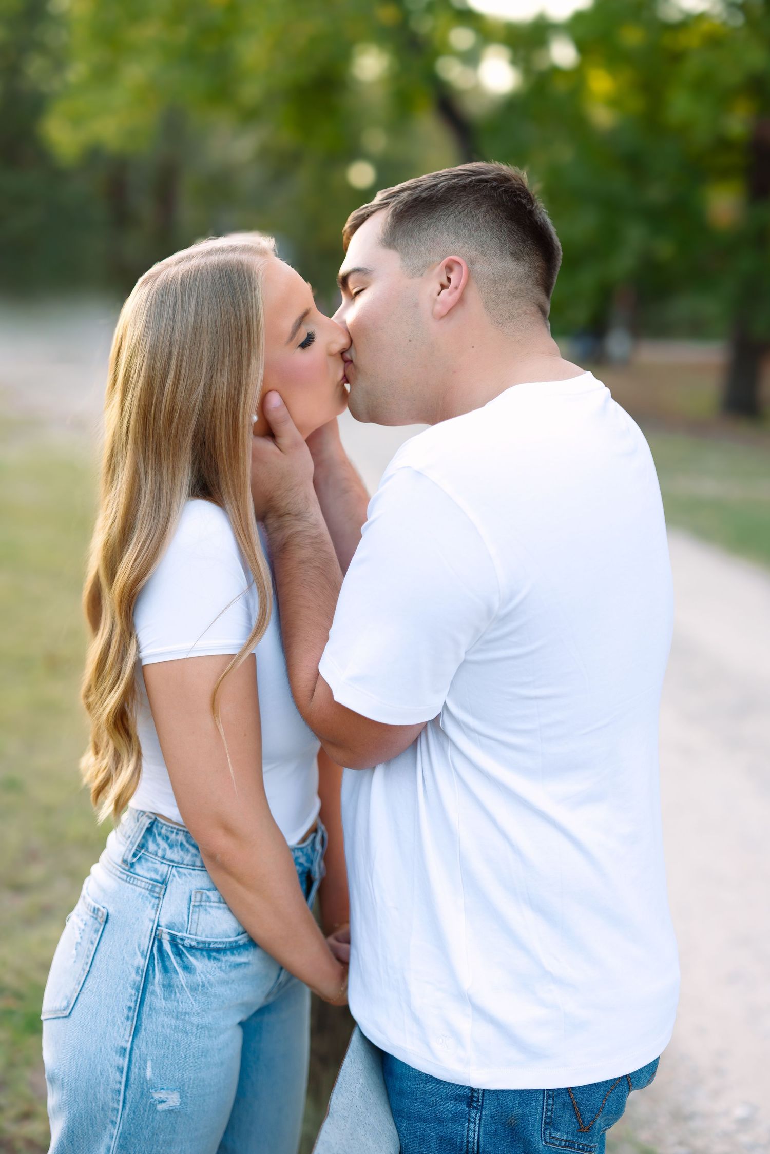 Couple kissing on a tree-lined path, both wearing white tops and jeans in a park setting.