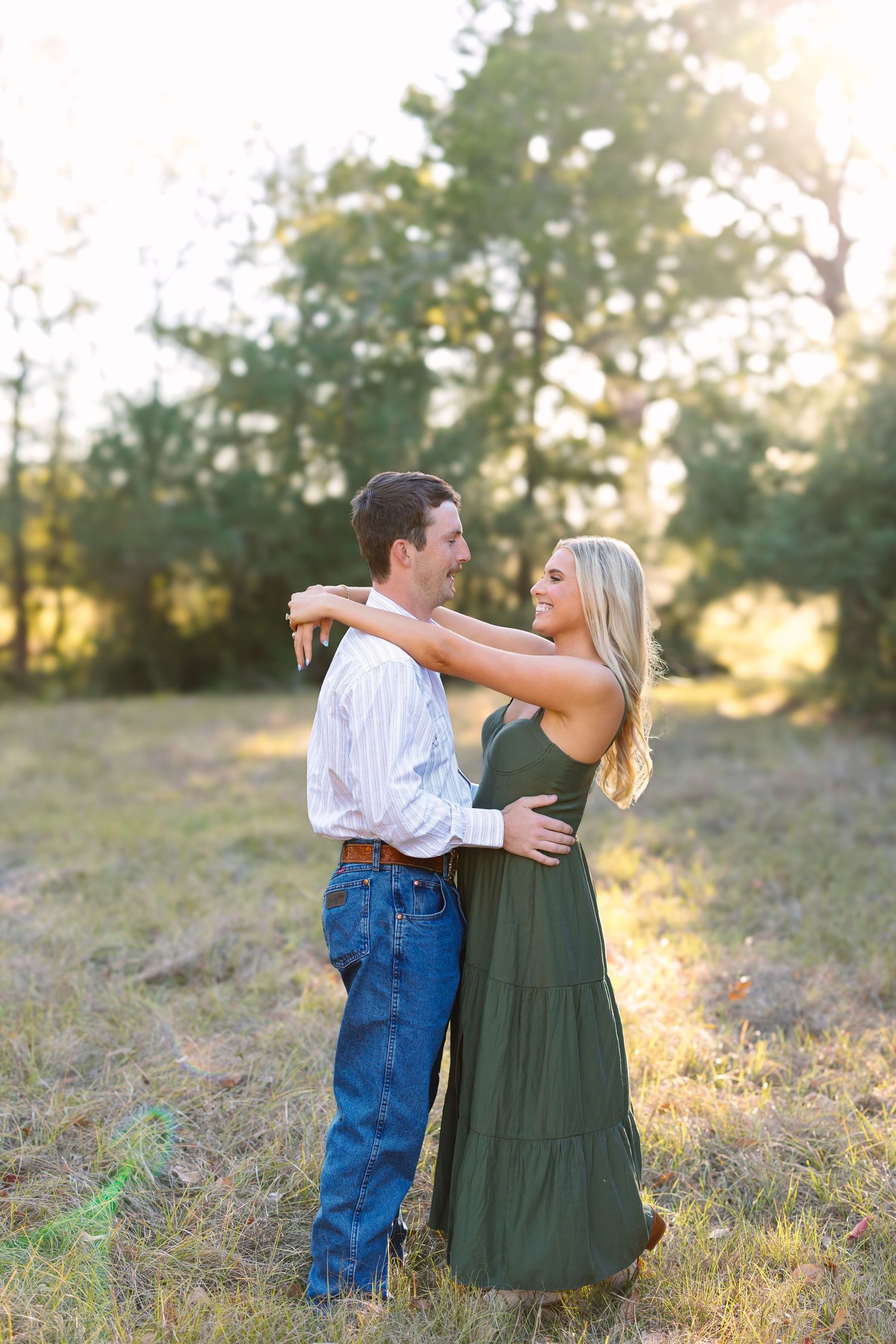 Couple embracing in sunlit field, woman in green maxi dress and man in white shirt and jeans smiling at each other.