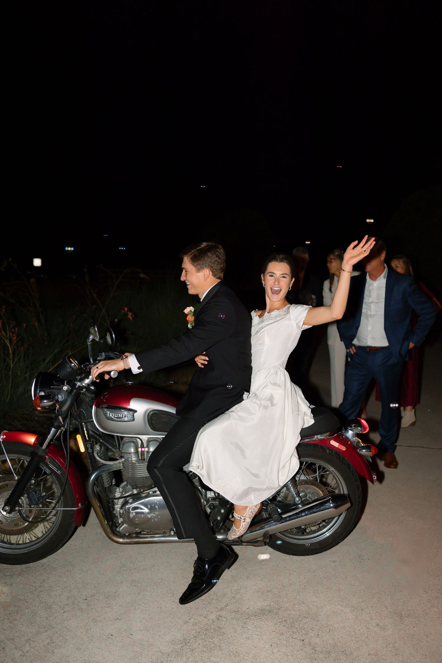 Excited bride and groom making a dramatic exit on a red motorcycle at night after their wedding.