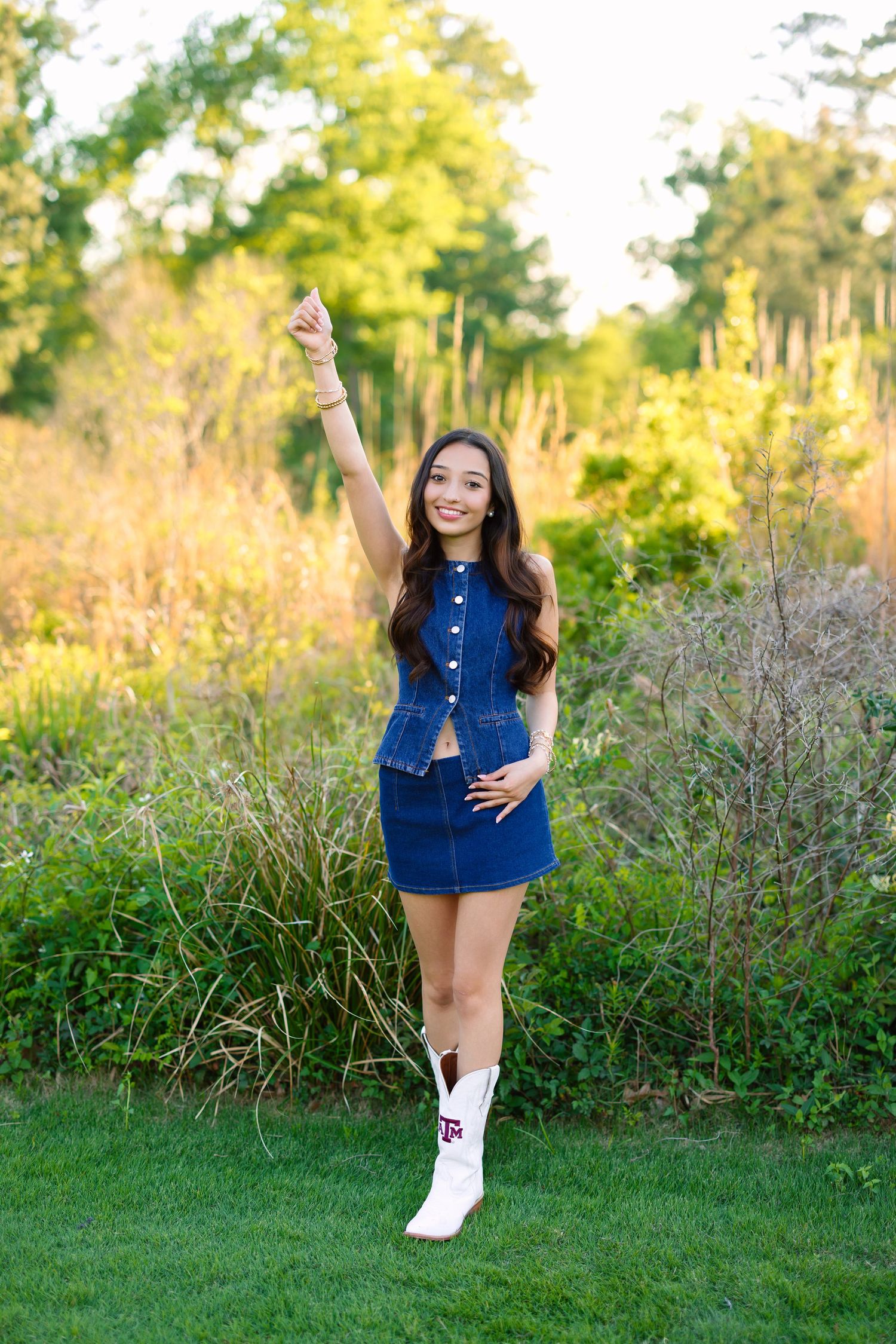 Young woman in denim skirt set and white cowboy boots poses outdoors pointing up, smiling in a lush green garden.