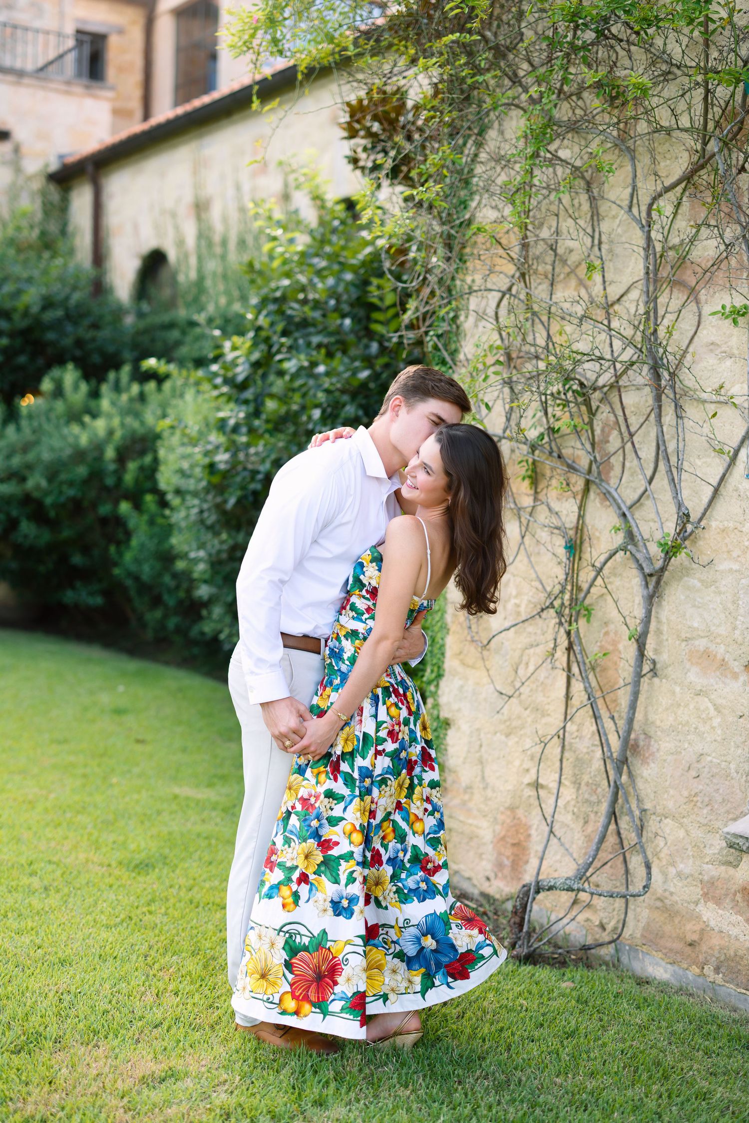 Couple embracing by a rustic stone wall in a garden, woman in floral dress, man in white shirt.