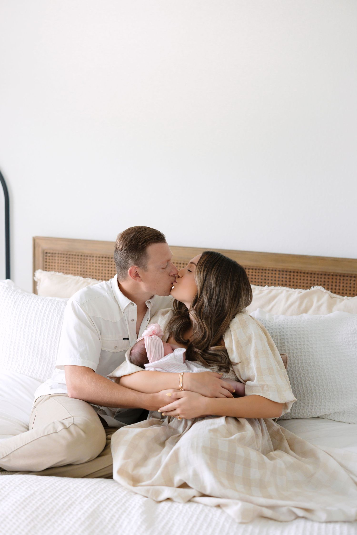 A couple shares a kiss on a bed while cradling their newborn baby, dressed in neutral tones.