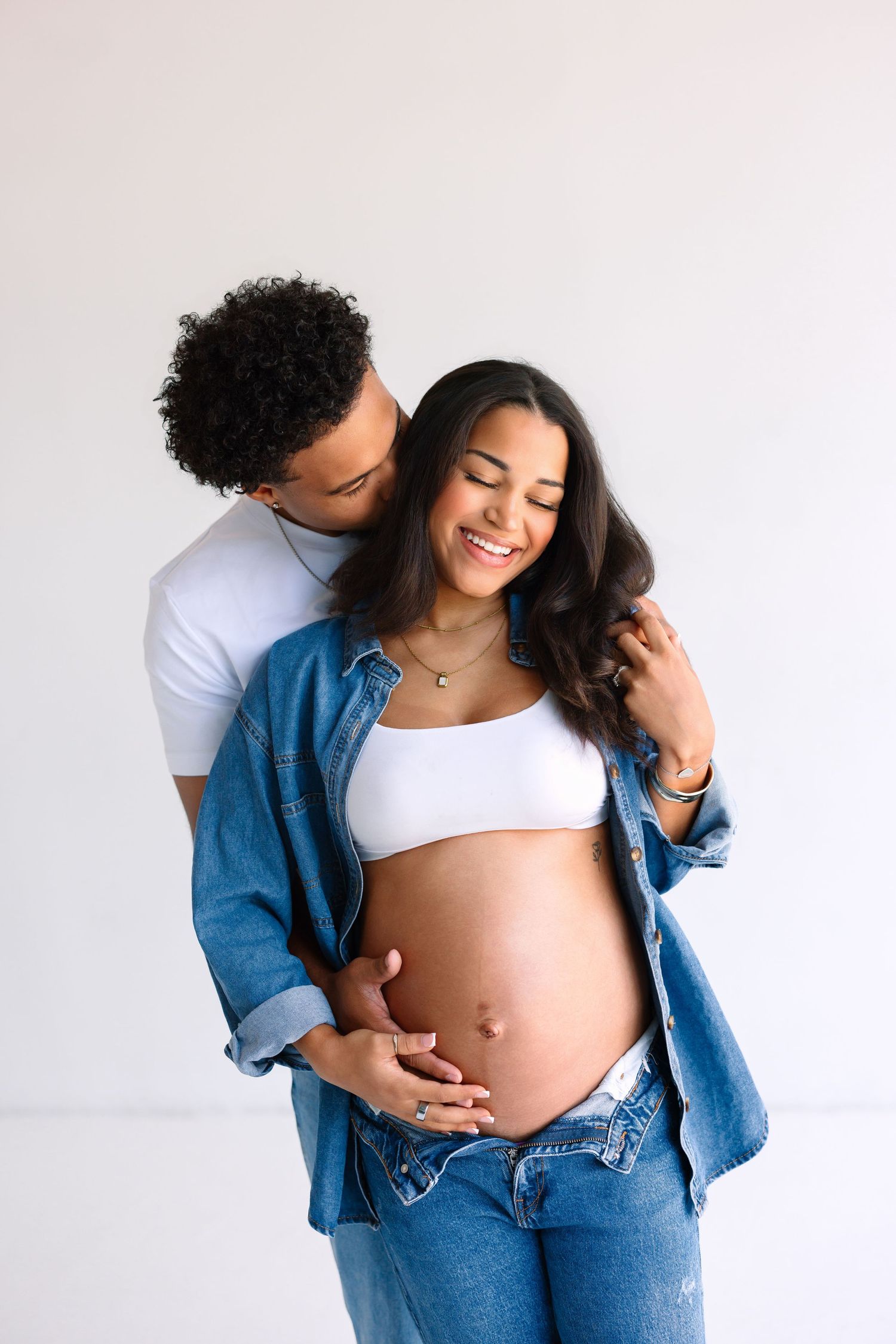 A couple poses for a maternity photo, with the man embracing the pregnant woman from behind against a white background.