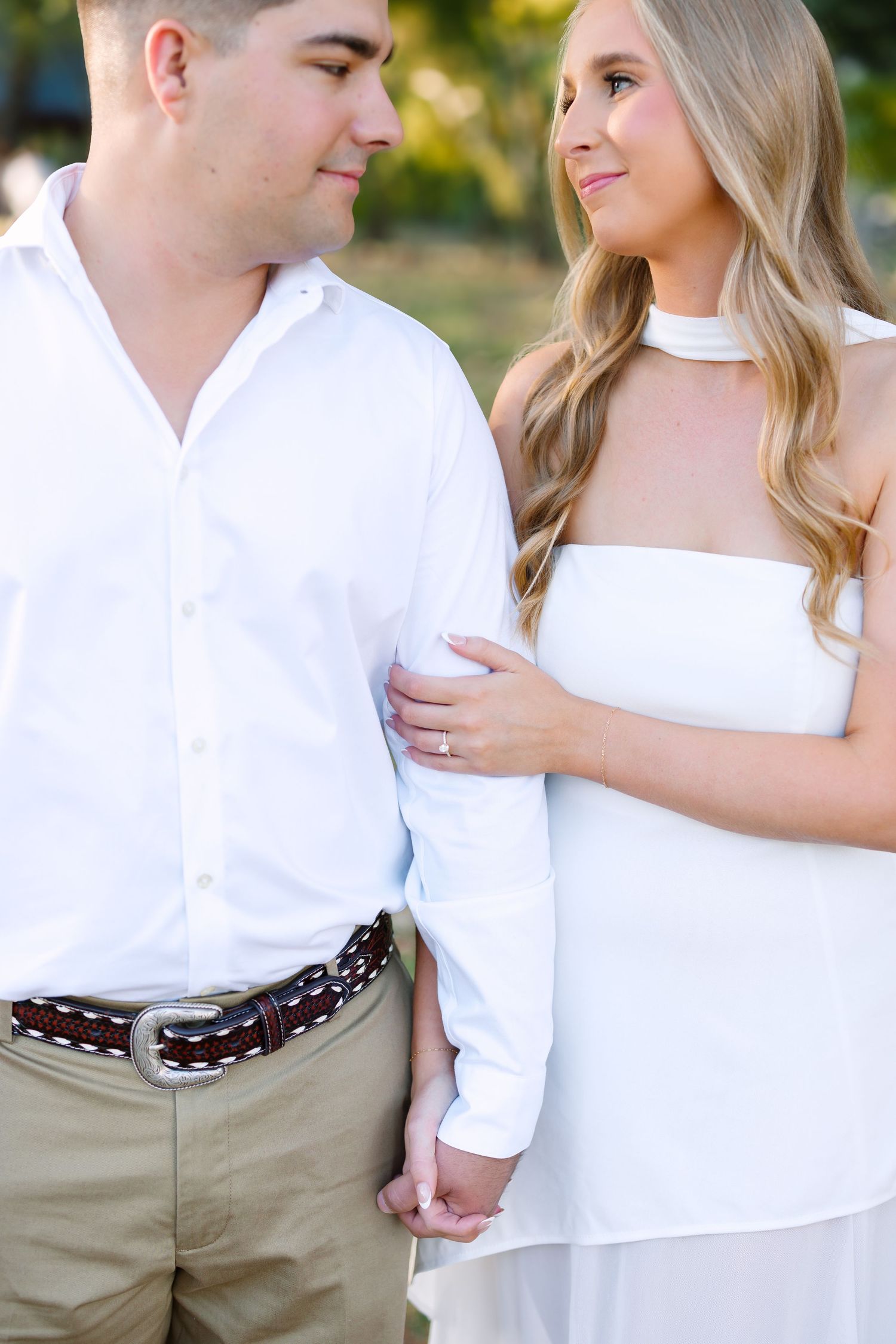 Couple in white outfits holding hands outdoors, woman wearing engagement ring smiles at man in white shirt and khakis.