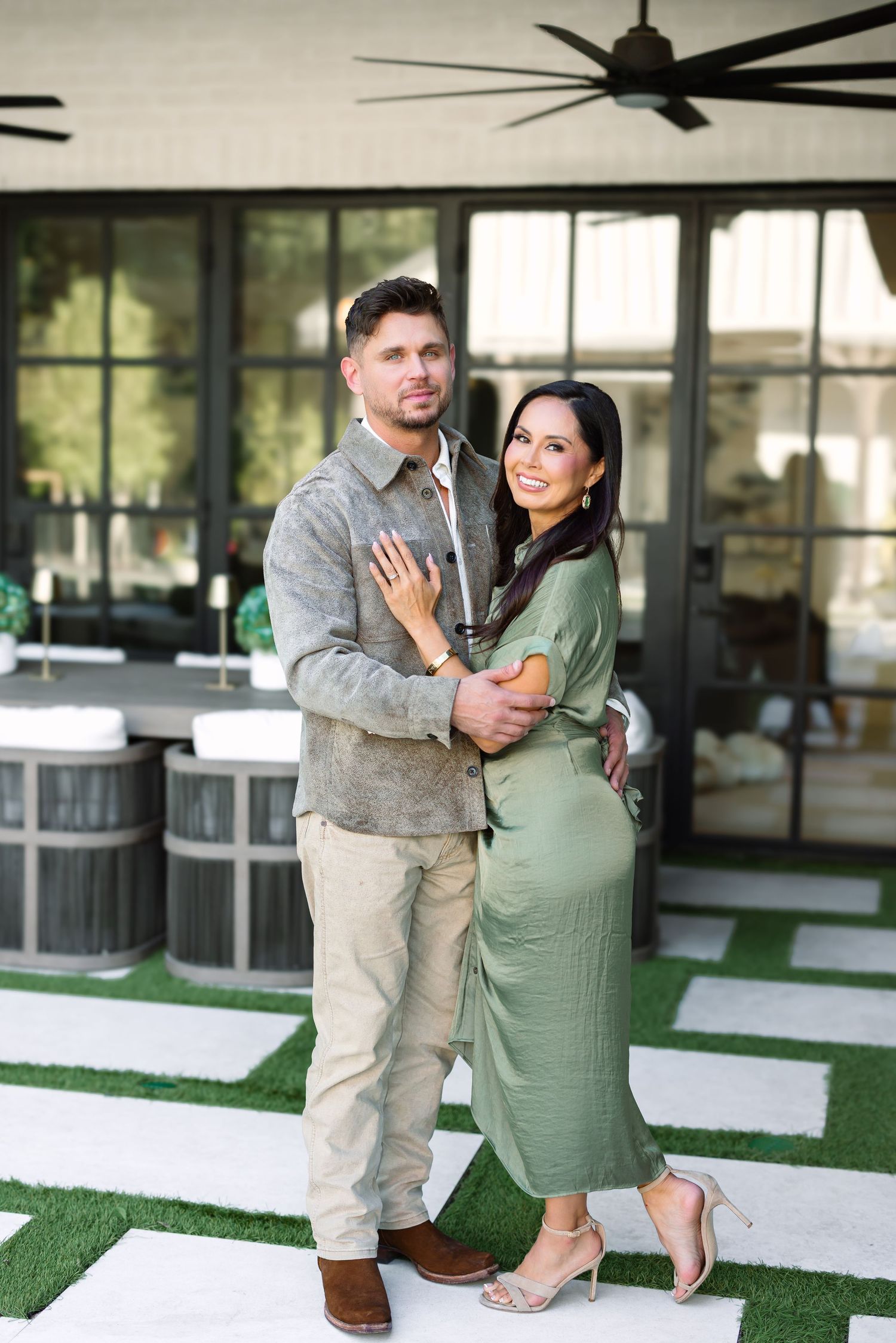 Couple posing outdoors, woman in green dress embracing man in grey jacket, modern patio with ceiling fan backdrop.