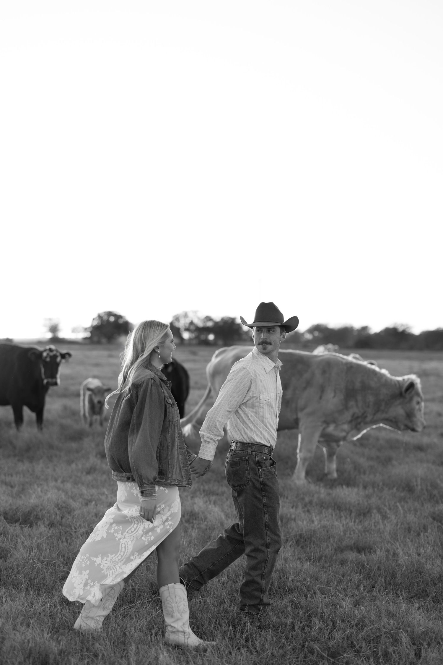 Black and white photo of a couple holding hands in a field with cattle, man wearing cowboy hat and woman in boots.