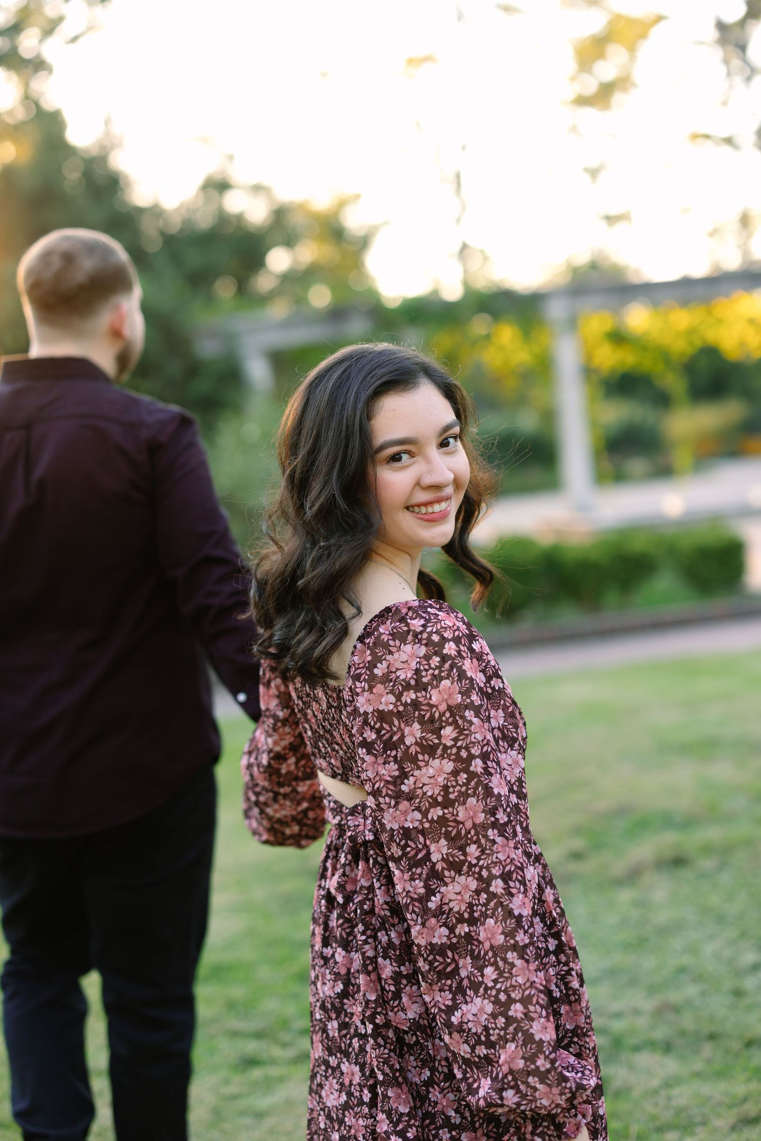 Smiling brunette woman in floral dress glances back while holding hands with her partner during a sunset garden photoshoot.