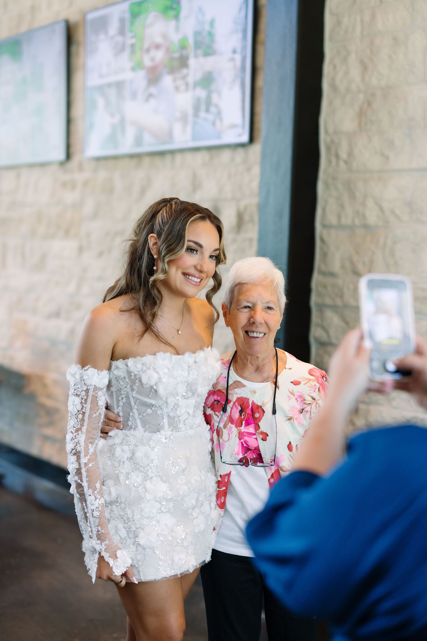 Bride in white lace mini dress poses with elderly woman in floral top while someone photographs them.