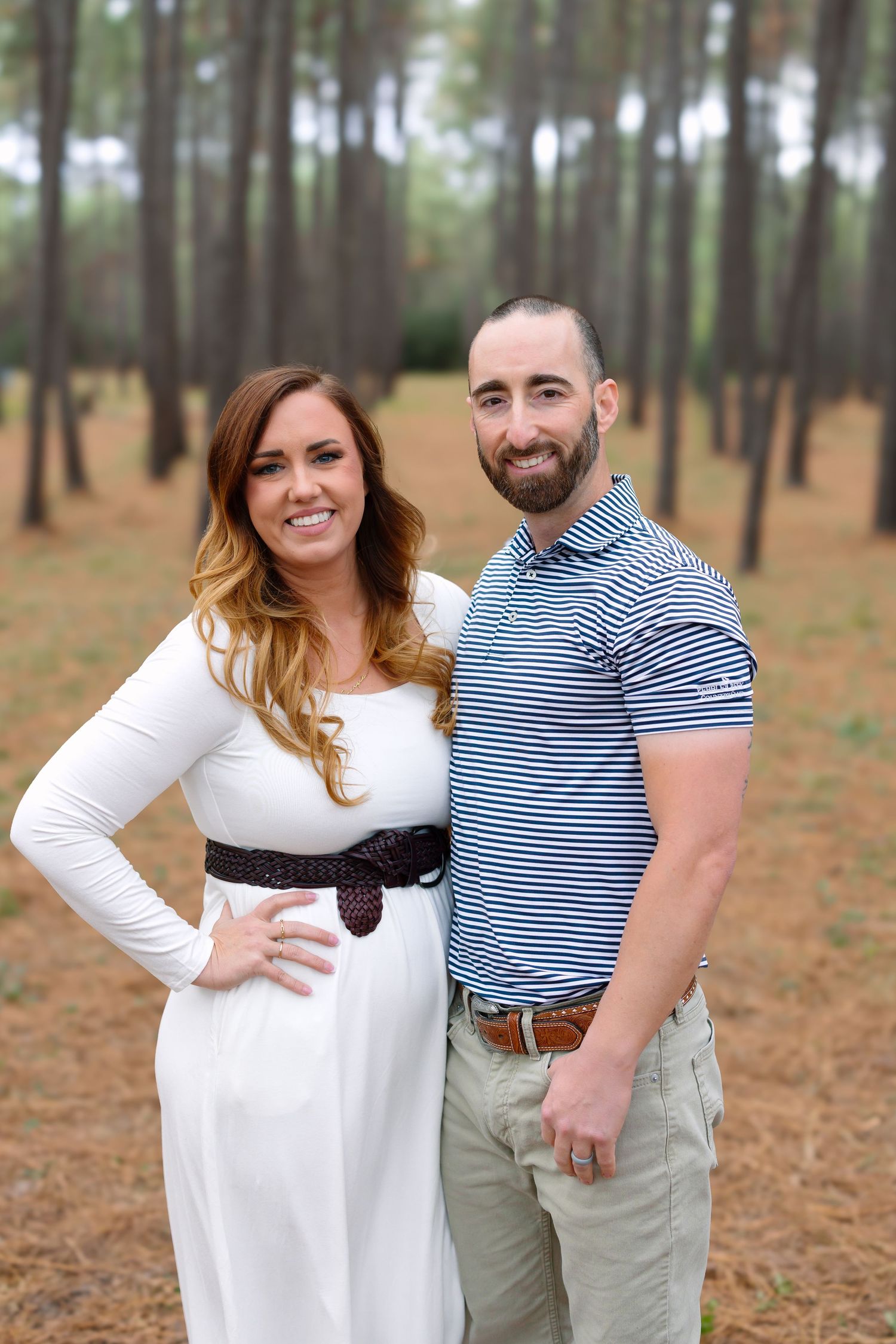 A smiling couple poses outdoors in a pine forest, woman in white dress with belt, man in blue striped polo shirt.