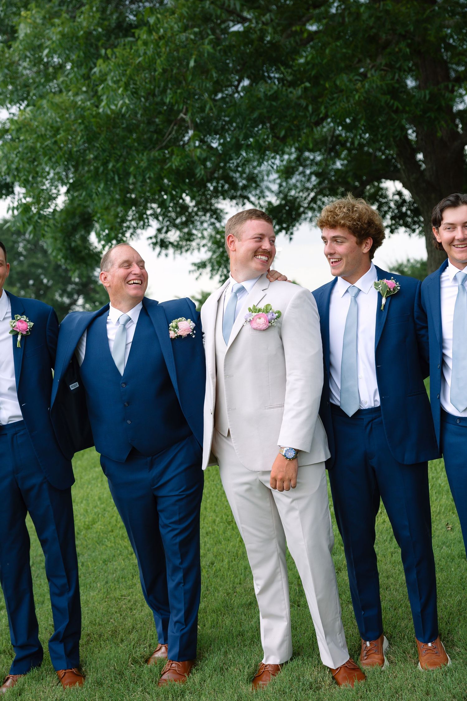 Groom in white suit stands with groomsmen in navy blue suits and boutonnieres, laughing outdoors on green grass.