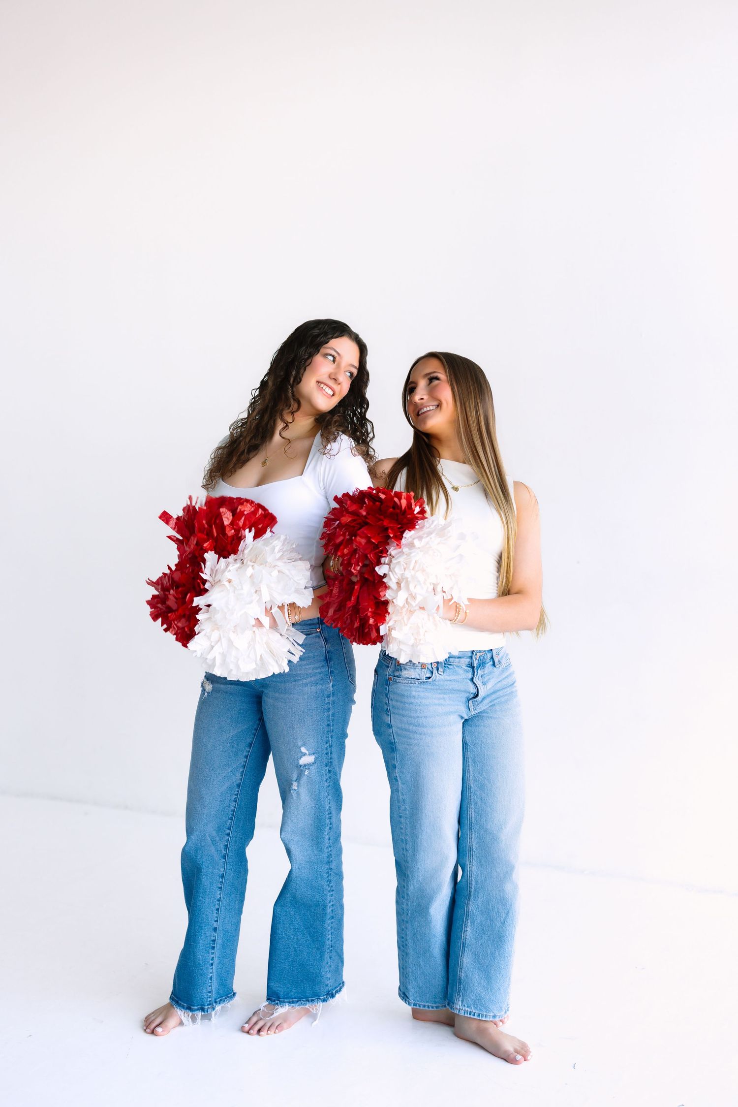 Women in jeans and white tops posing with red pom poms against white background.