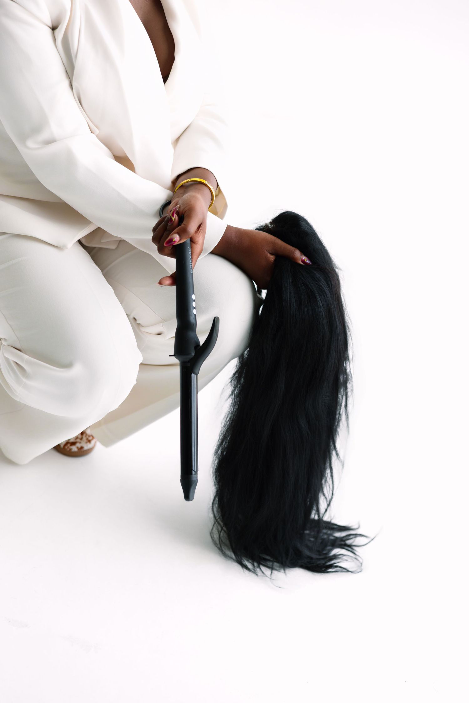 Woman in white suit using a curling iron on long black hair against a white background.